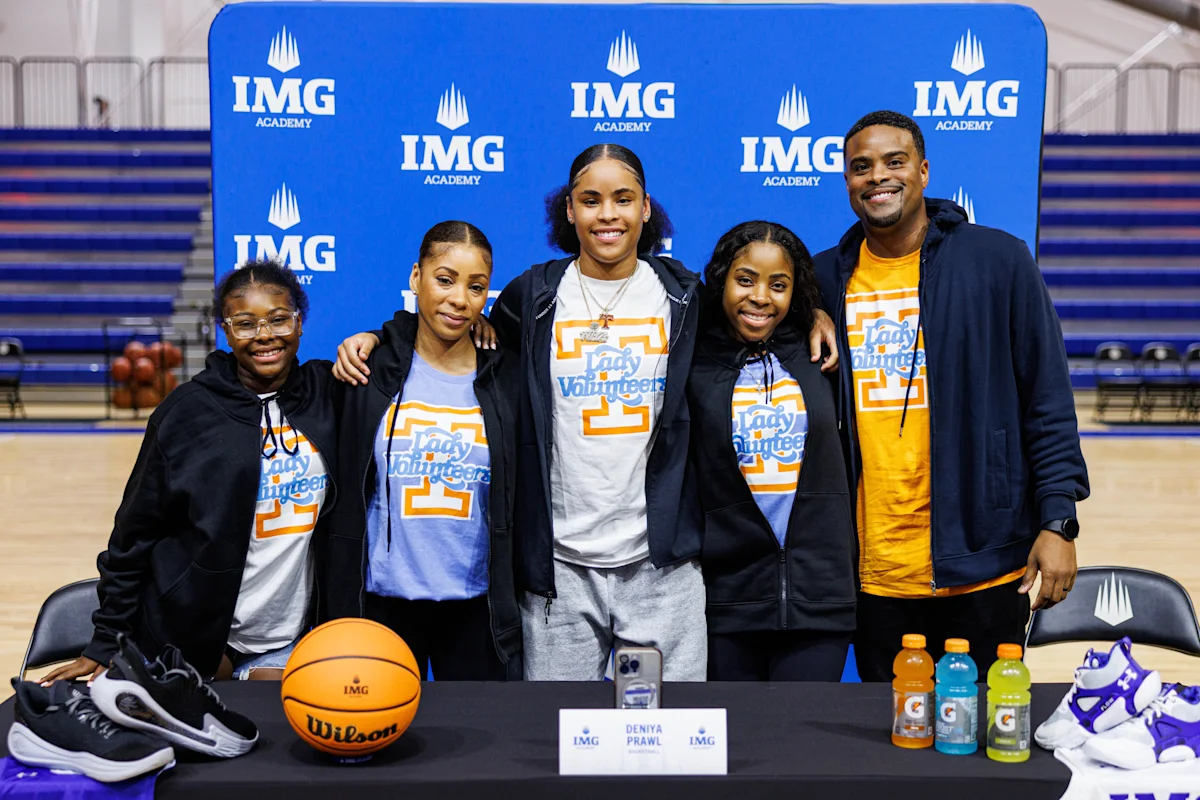 IMG Academy basketball player stands with family on signing day, wearing a Tennessee shirt behind a table with a ball, shoes, and drinks, celebrating her college commitment.
