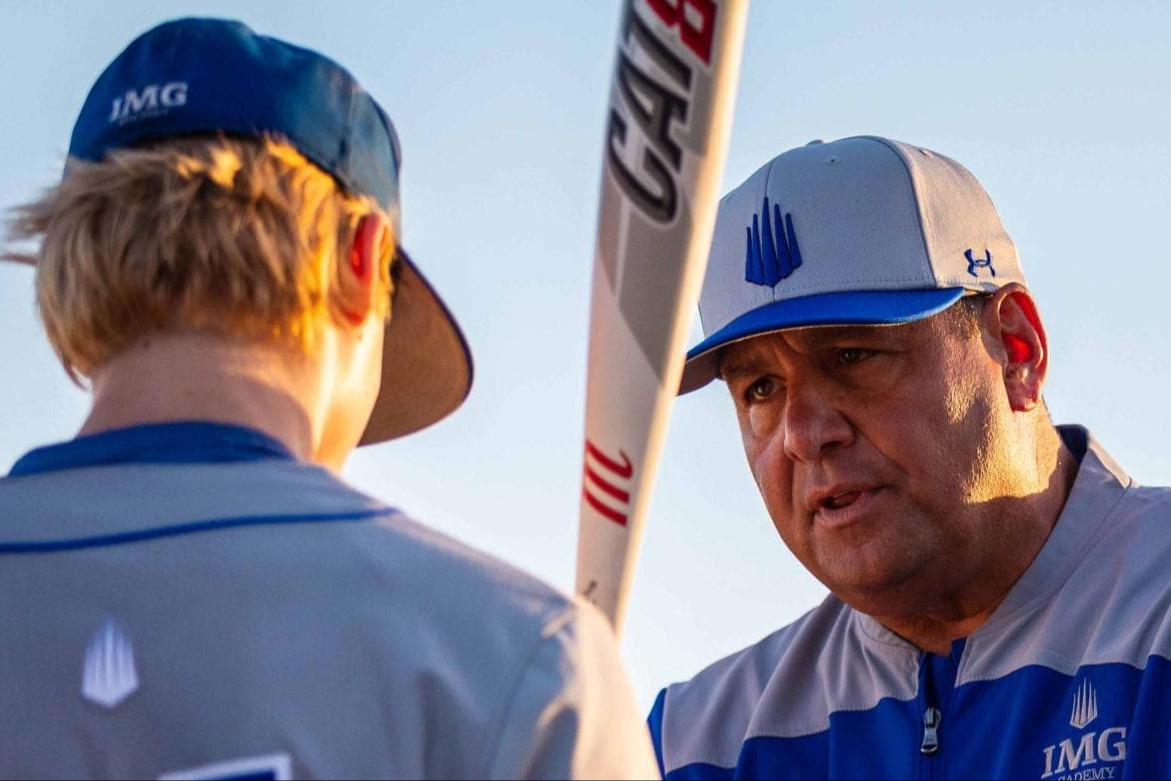 An IMG Academy baseball coach speaks intently to a young athlete during practice, holding a bat vertically as they discuss technique as the sun sets in the background.