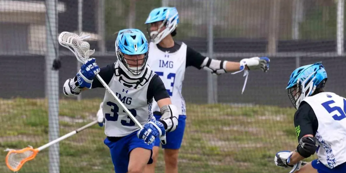 Helmeted lacrosse players compete near a goal.