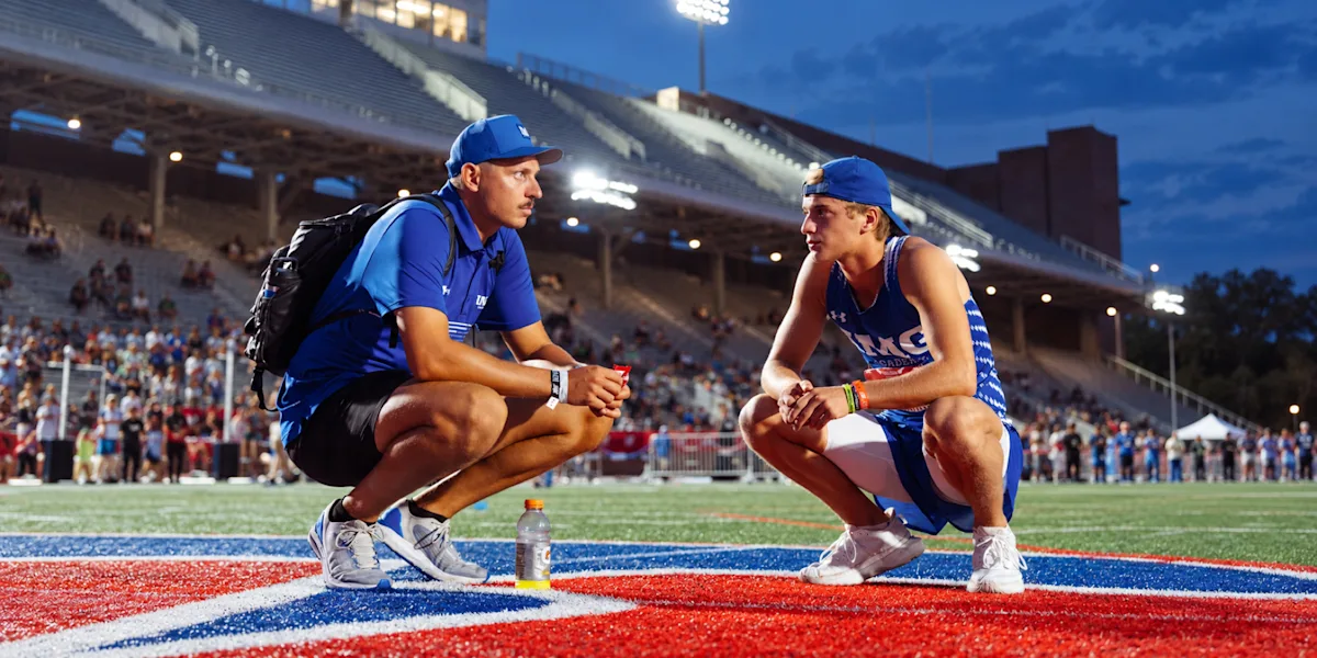 Two track athletes crouch together and talk beside a track.