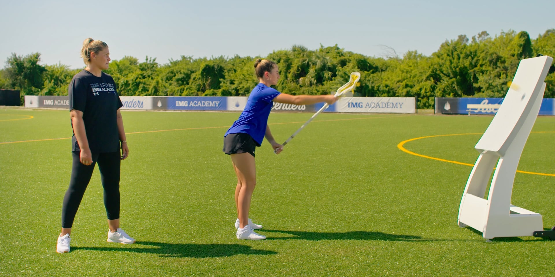 A lacrosse player practices a draw toward a training target while a coach observes.