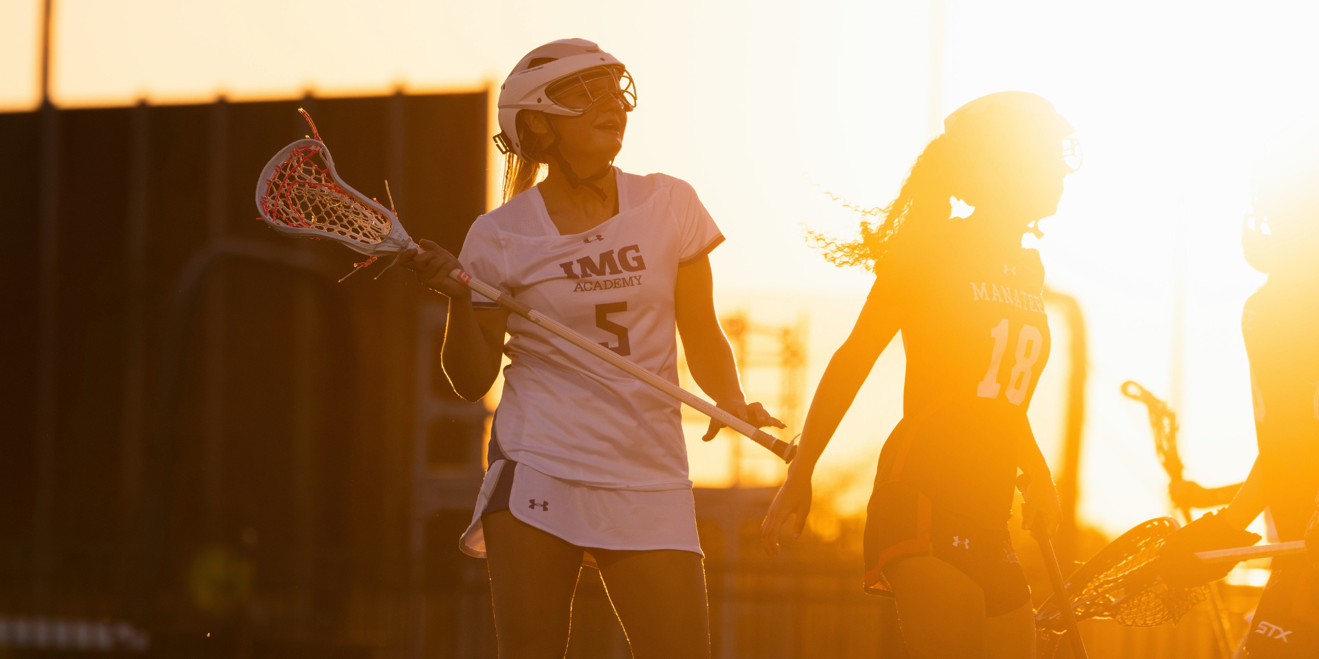 Lacrosse players stand together on the field in warm evening sunlight.