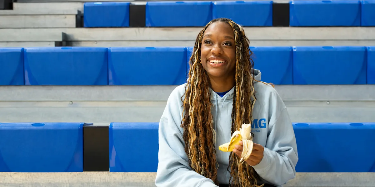 An athlete smiles while holding a peeled banana on gym bleachers.