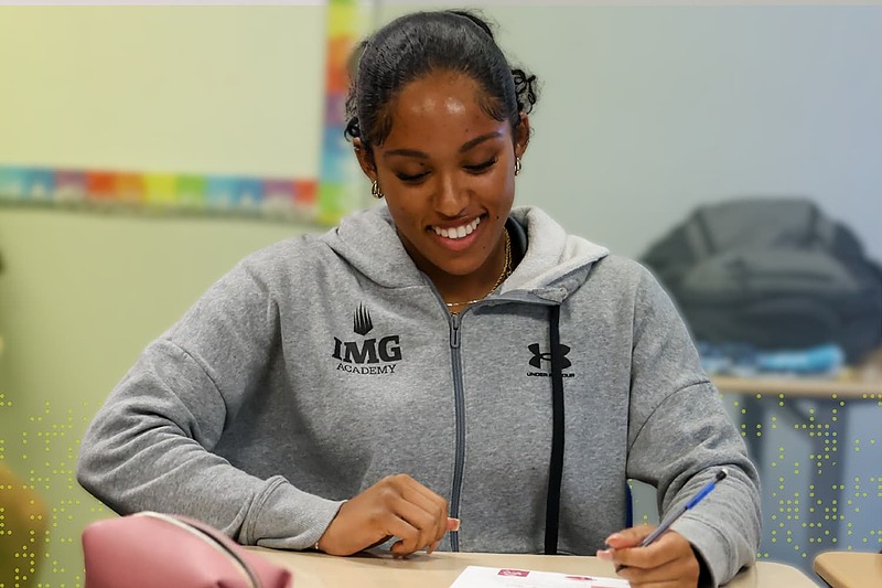 IMG Academy student-athlete wearing a gray IMG Academy hoodie smiles while writing at a desk in a classroom setting.