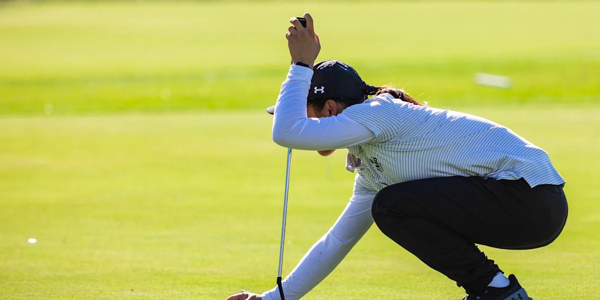 A golfer crouches on a putting green while using a putter for support.