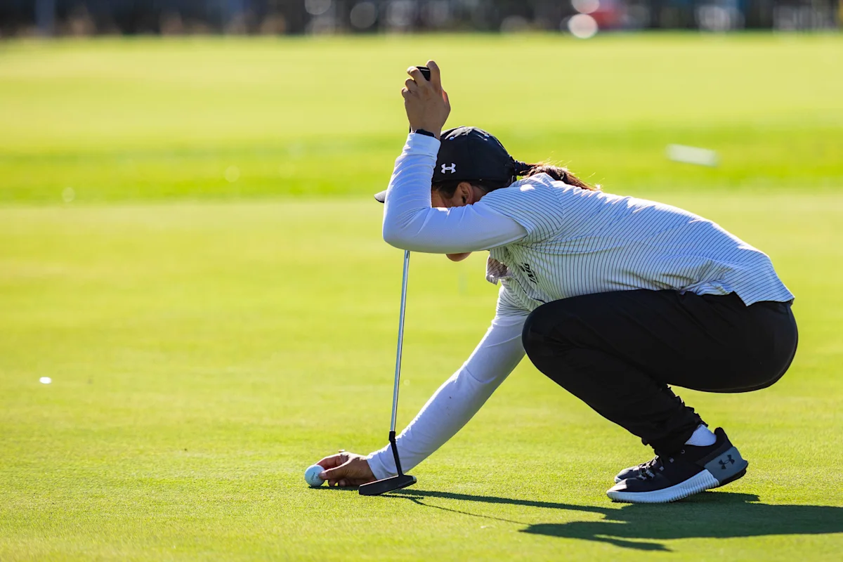 Golf student-athlete wearing a long sleeve polo, hat, and pants crouches to align a putt during practice, showcasing concentration and visualization techniques on the green.