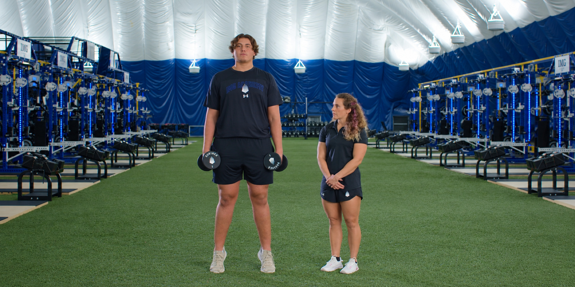 An athlete holds dumbbells at his sides during a calf-raise exercise beside a trainer.