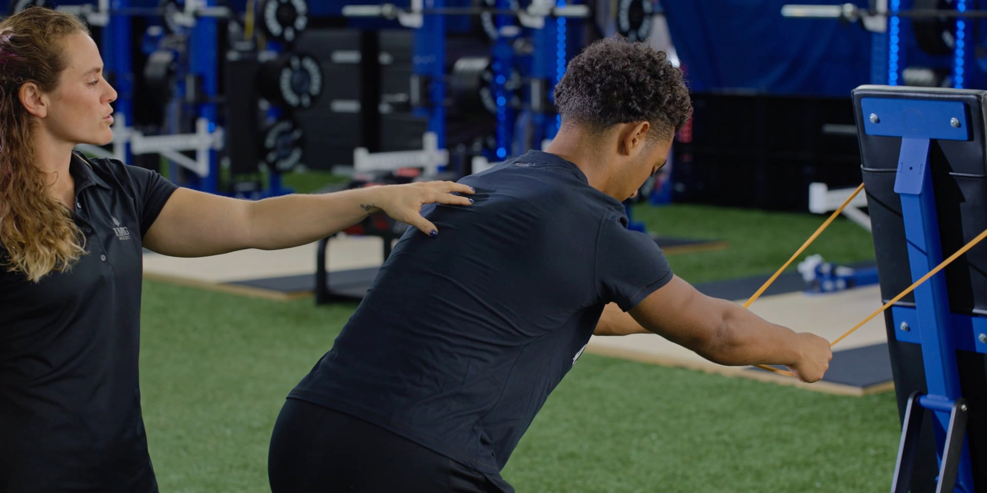 A trainer guides an athlete performing a resistance-band exercise attached to a rack.
