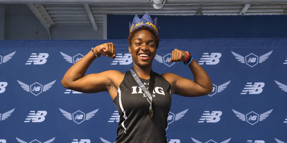A smiling athlete flexes both arms in front of a sports-event backdrop.