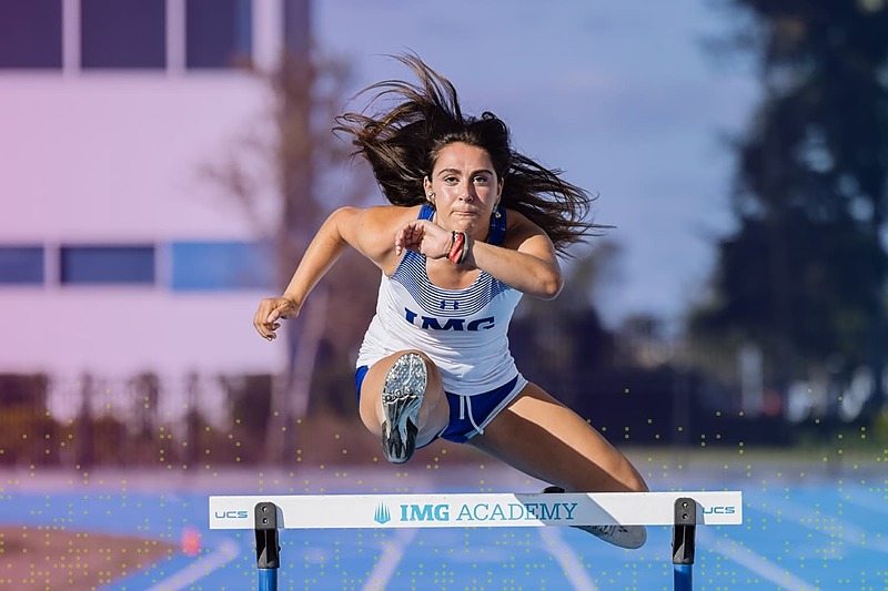 IMG Academy track and field athlete clears a hurdle during a sprint hurdle event, captured midair with strong form and focus on an outdoor track.