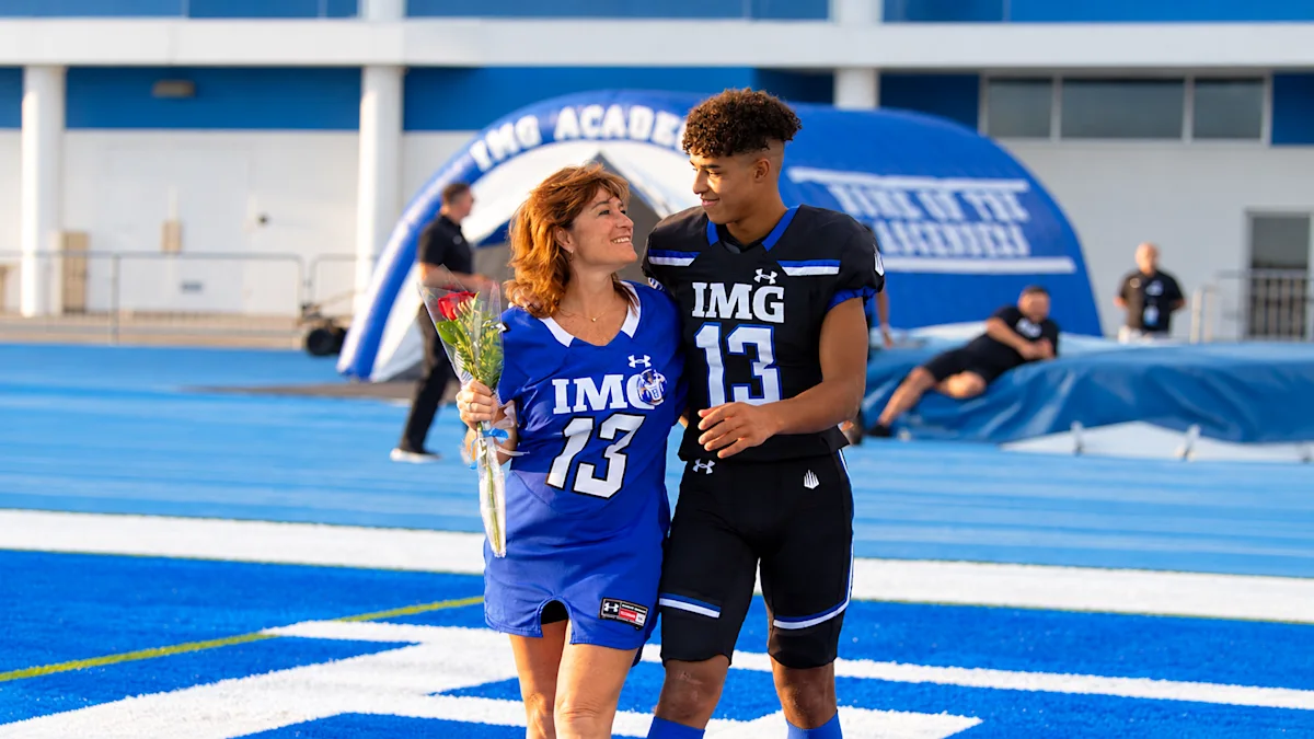 Youth football player in a black and blue IMG Academy uniform standing with his mother on the blue turf during Senior Night or Parent's Weekend.
