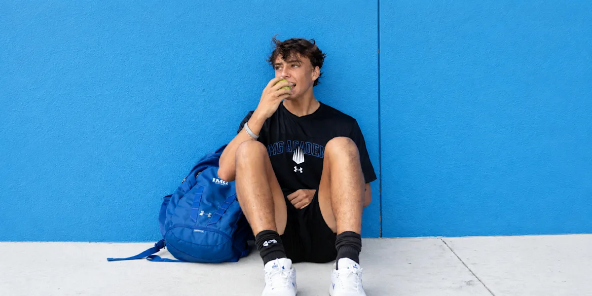 An athlete sits against a blue wall eating a snack beside a backpack.