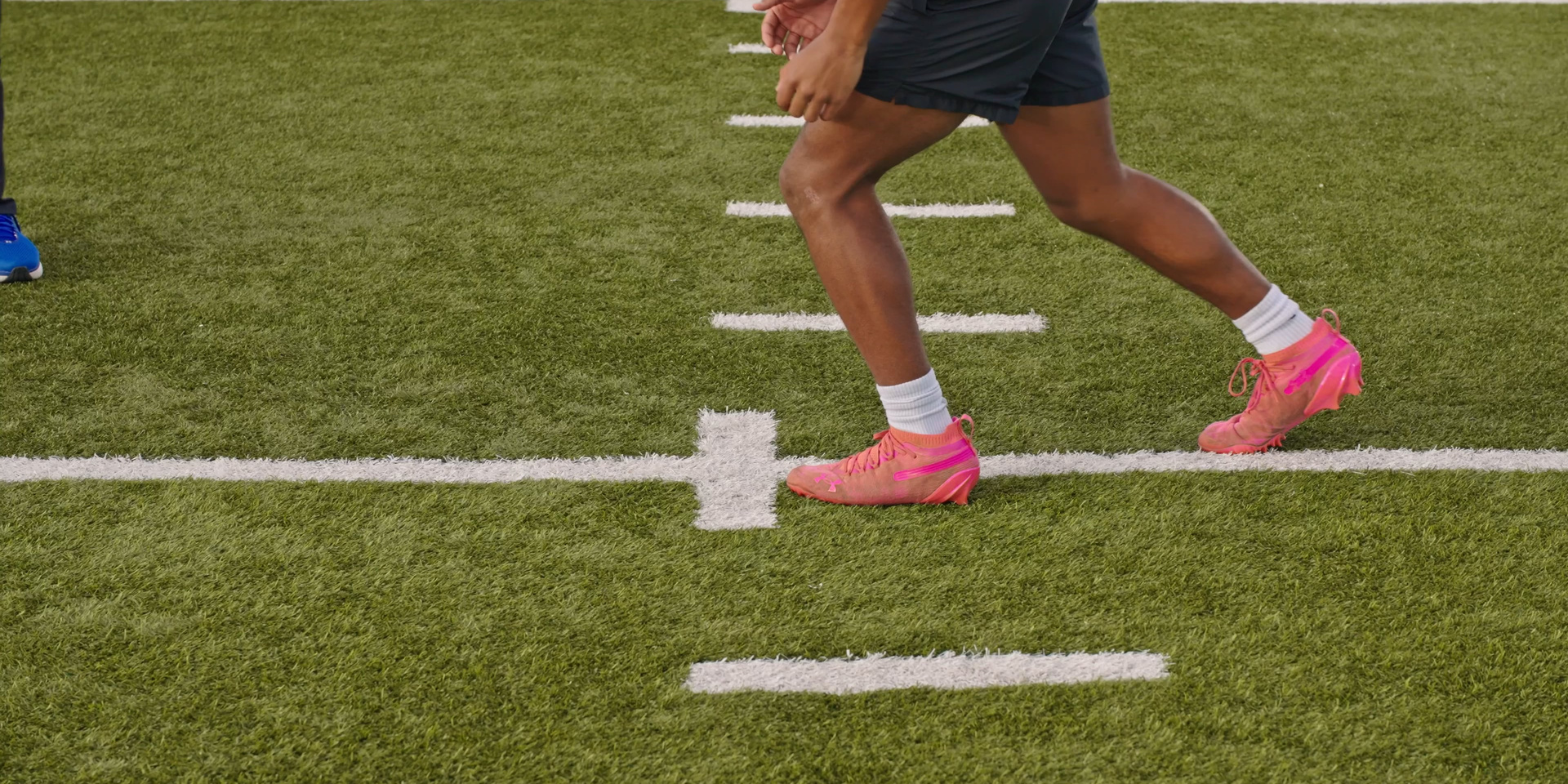 A close view shows an athlete’s legs and pink cleats moving over field markings.