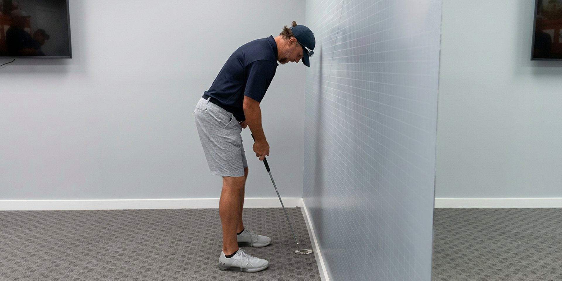 A golfer demonstrates a putting posture beside an indoor wall.