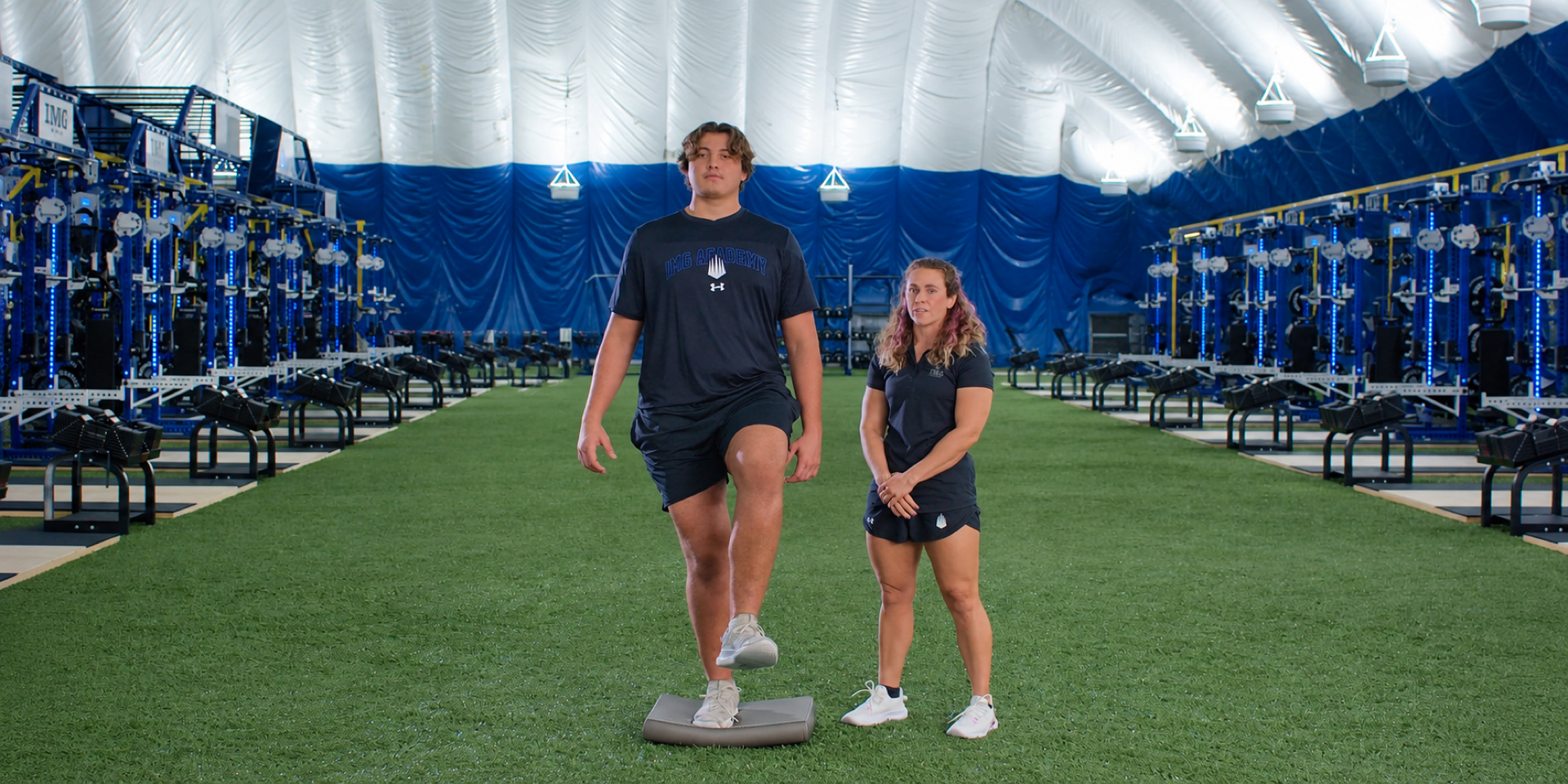 An athlete balances on one leg on a padded surface beside a trainer.
