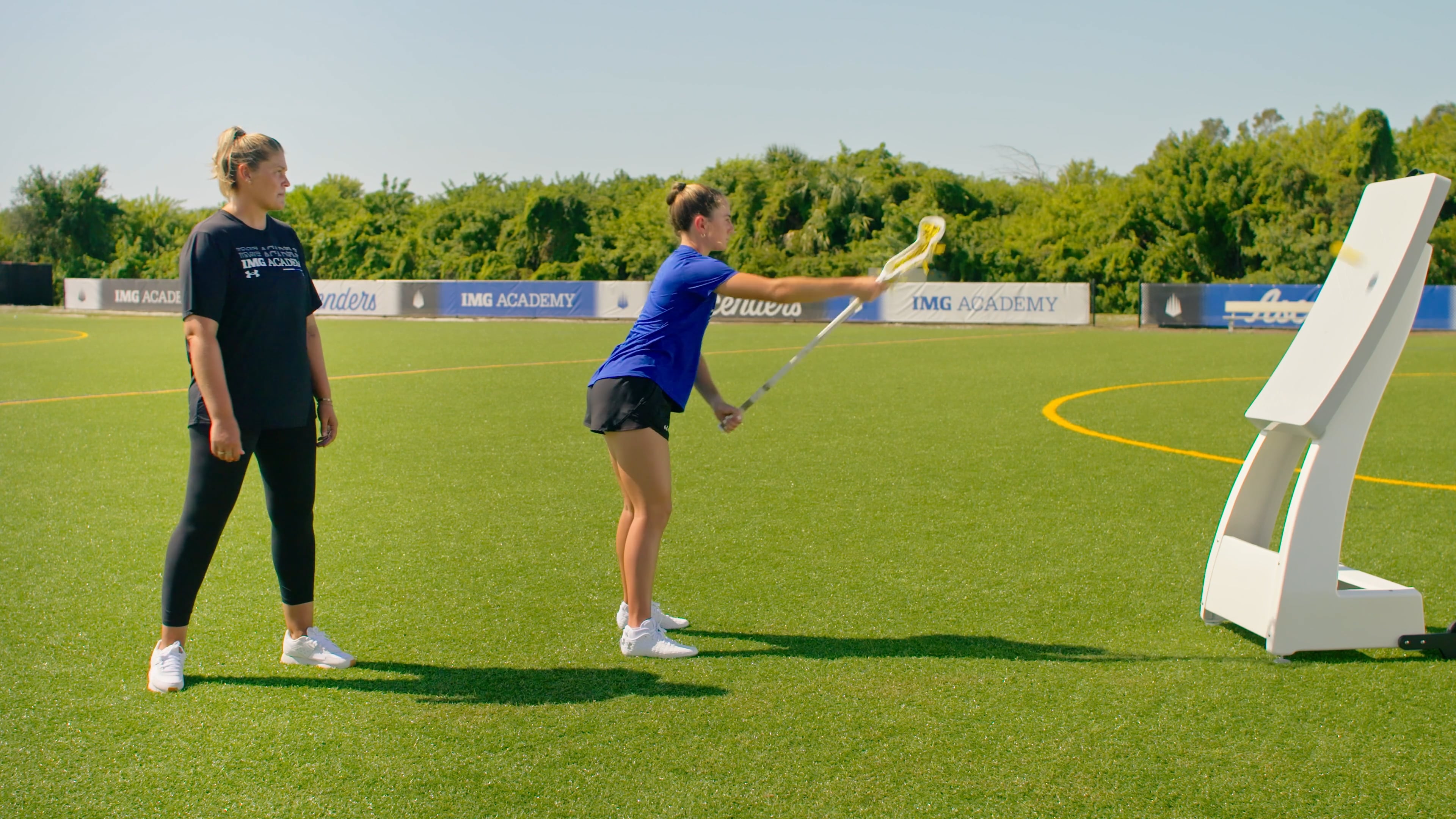 A female lacrosse player practices throwing and catching using a white rebounder screen on a turf field, while a coach stands behind her observing her form.