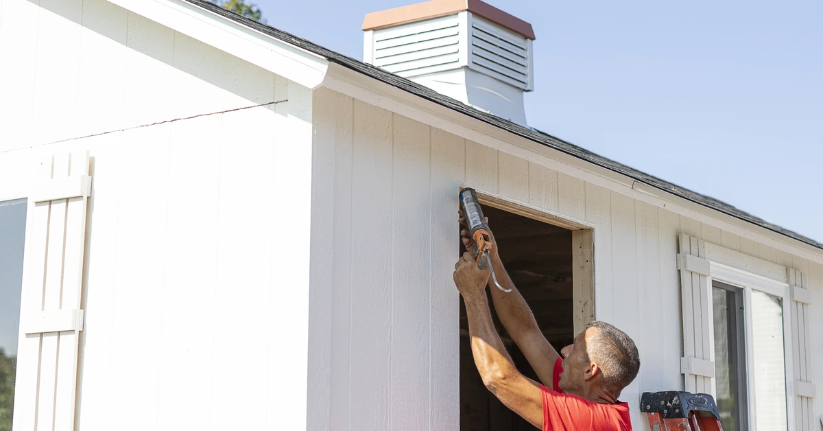 man in red shirt applying sealant to roof of shed with white siding