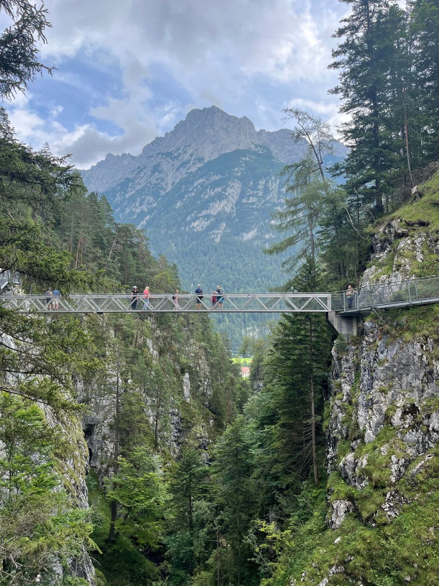 Panoramiczny most Panorama-Brücke w wąwozie Leutascher Geisterklamm. Na moście pozują turyści. W tle panorama Alp.