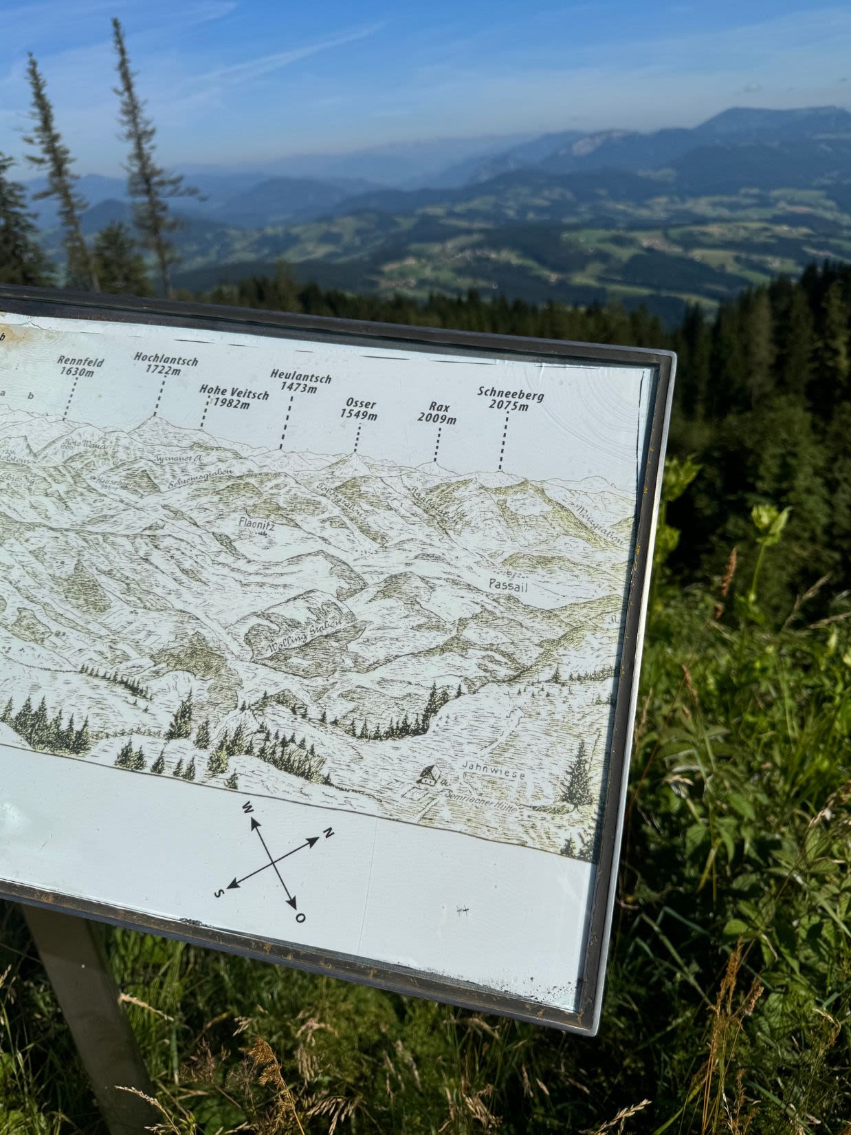 Tablica pokazująca panoramę gór, którą widać ze szczytu Schöckl, stoi wśród zieleni. Na horyzoncie pola i lasy, brak zabudowań.