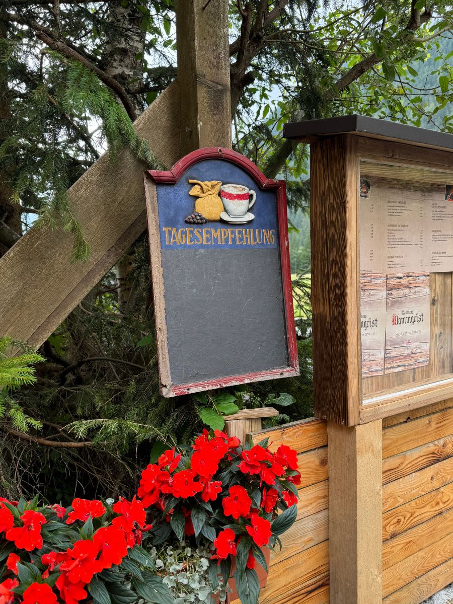 Wejście do restauracji Gasthaus Klammgeist w Austrii. Obok drewnianej tablicy z ogłoszeniami stoją doniczki z czerwonymi kwiatami.
