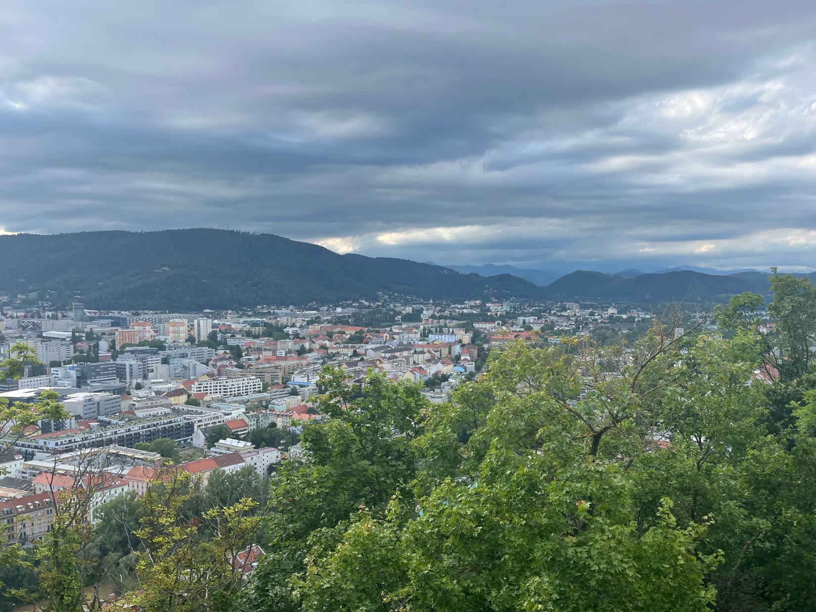 Widok na Graz ze wzgórza zamkowego Schlossberg. Na pierwszym planie drzewa liściaste, w dole panorama na miasto, dalej niewielkie wzgórza. 