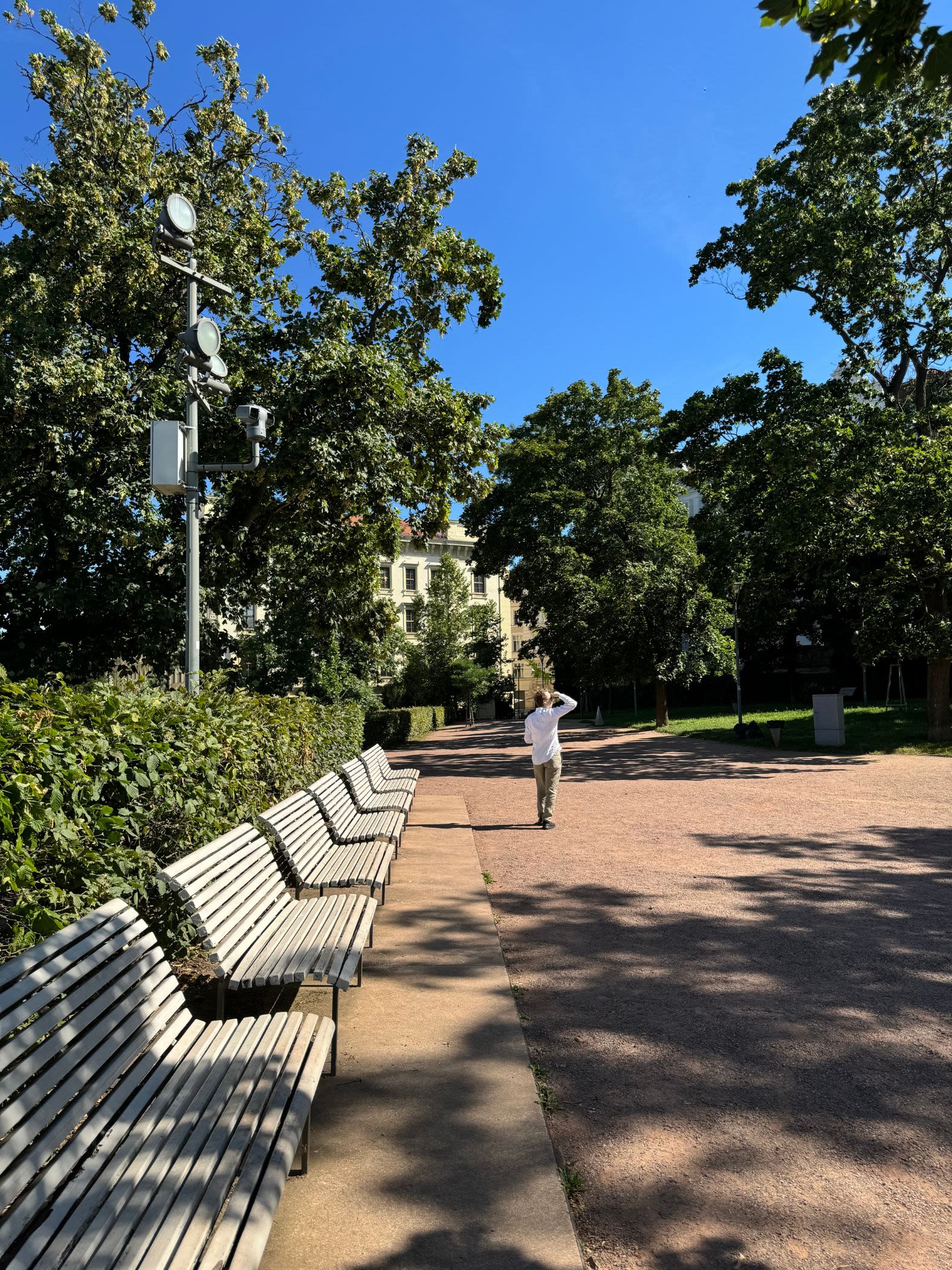 Park niedaleko punktu widokowego Vyhliadka Husova. Wzdłuż żwirowej drogi stoi rząd białych ławek. W parku jest dużo zieleni.