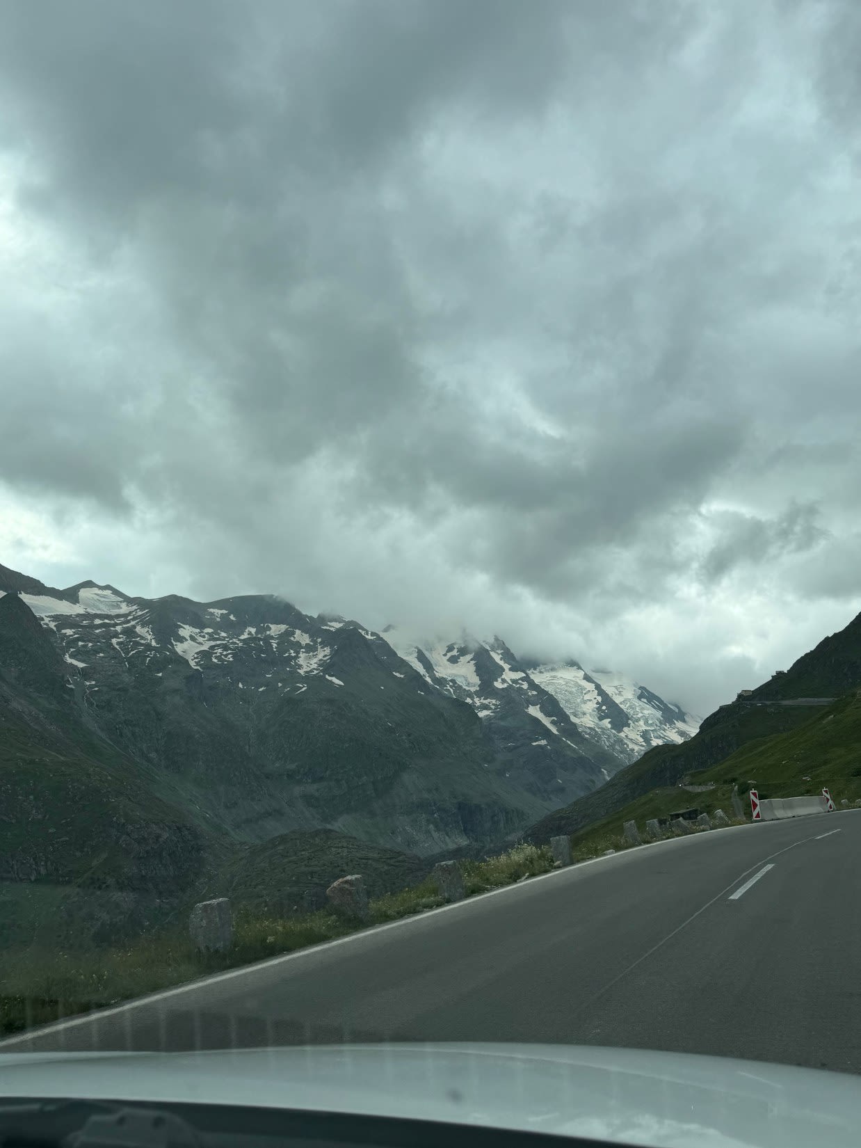 Samochód jedzie po alpejskiej drodze Grossglockner Hochalpenstrasse w Austrii w lipcu. Niebo pokryte jest chmurami, na zboczach Alp leży śnieg. 