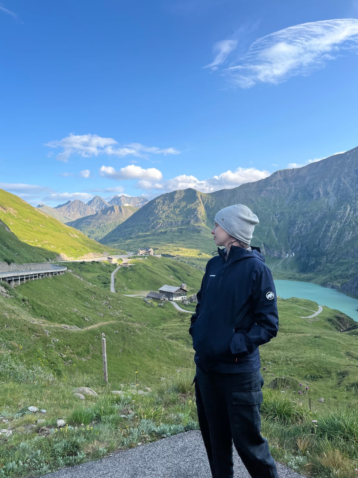 Autorka bloga Słowa Płyną patrzy na górski krajobraz na trasie Grossglockner Hochalpenstrasse. W oddali znajduje się droga. W dole górska zabudowa. 