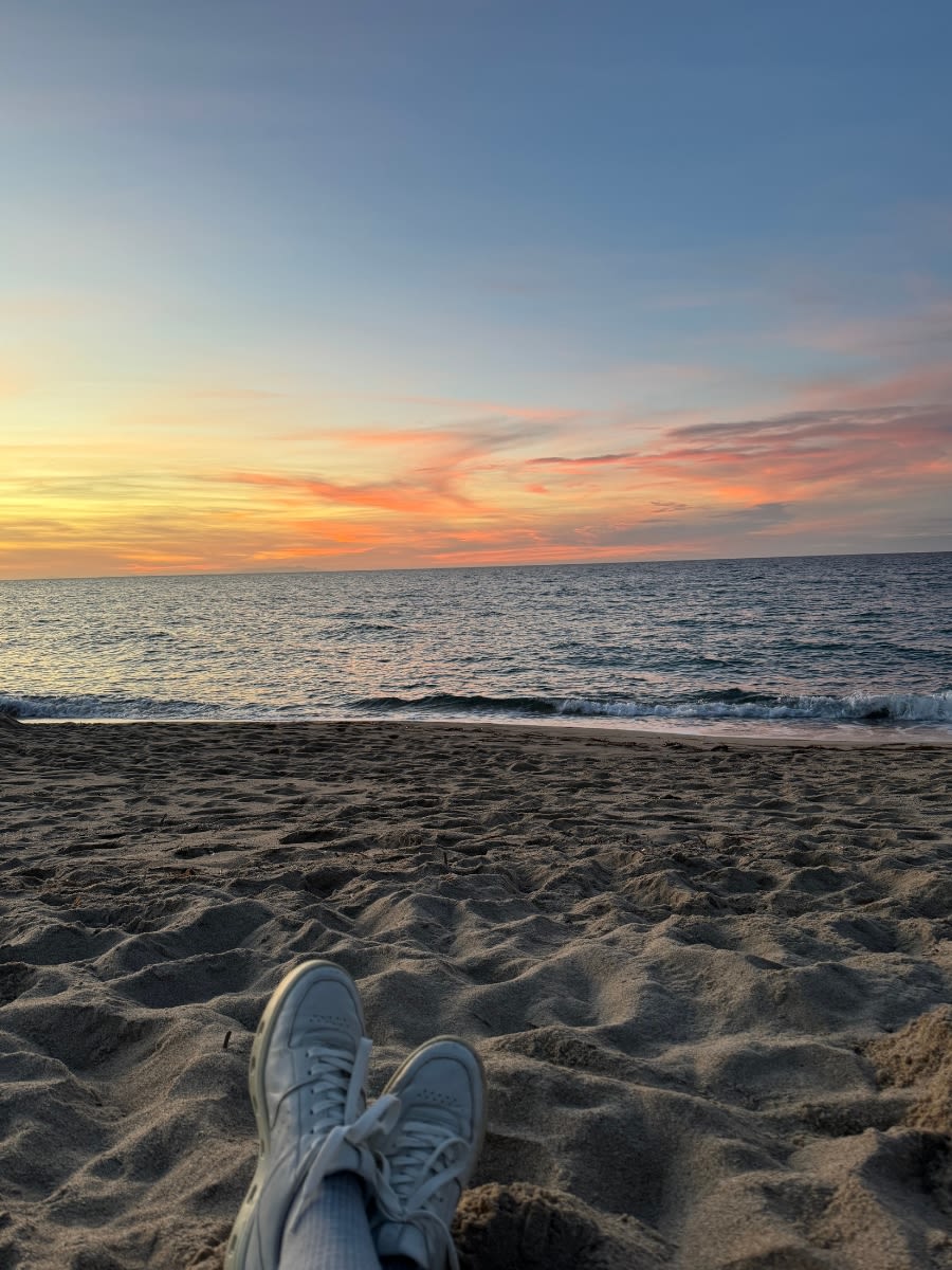 Plaża Spiaggia San Pietro A Mare di Valledoria o zachodzie słońca na Sardynii. Niebo jest w kolorach pomarańczu, różu i żółci. Woda jest spokojna. Osoba, która robi to zdjęcie leży na piasku i patrzy na morze.