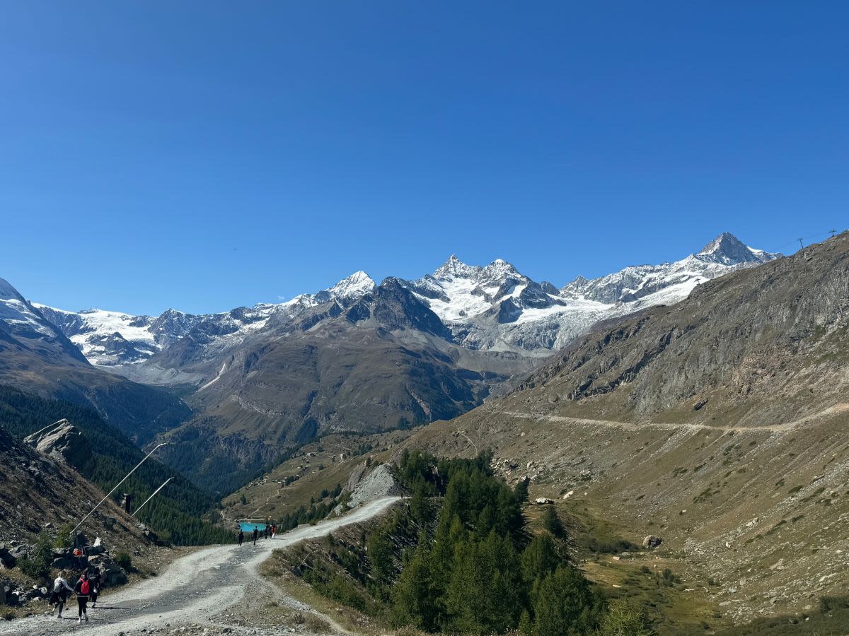 Szlak Pięciu Jezior, z którego można podziwiać Matterhorn. Trasa jest łatwa i przyjemna. Na ostatnim planie ośnieżone szczyty gór. Na pierwszym planie turyści.