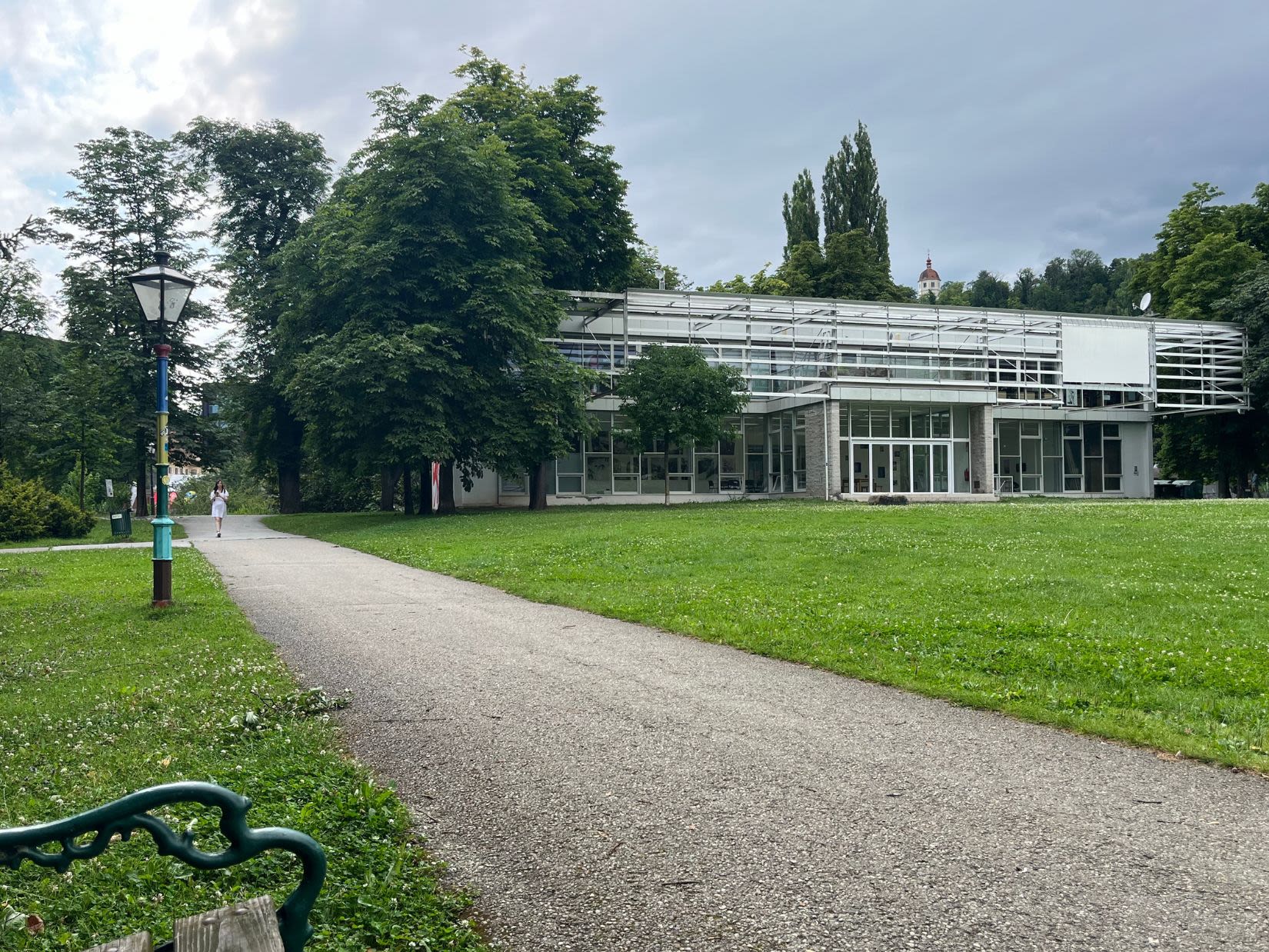 Park Stadtpark w Grazu w lecie. Alejka w parku jest żwirowana, a trawa równo przycięta. Znajduje się tu szklany budynek. 