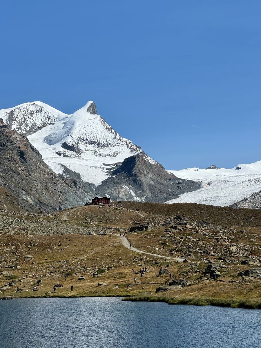 Alpejskie góry w słoneczny wrześniowy dzień. Na pierwszym planie jezioro, przy którym chodzą ludzie. Dalej ośnieżone szczyty górskie. Kanton Valais.