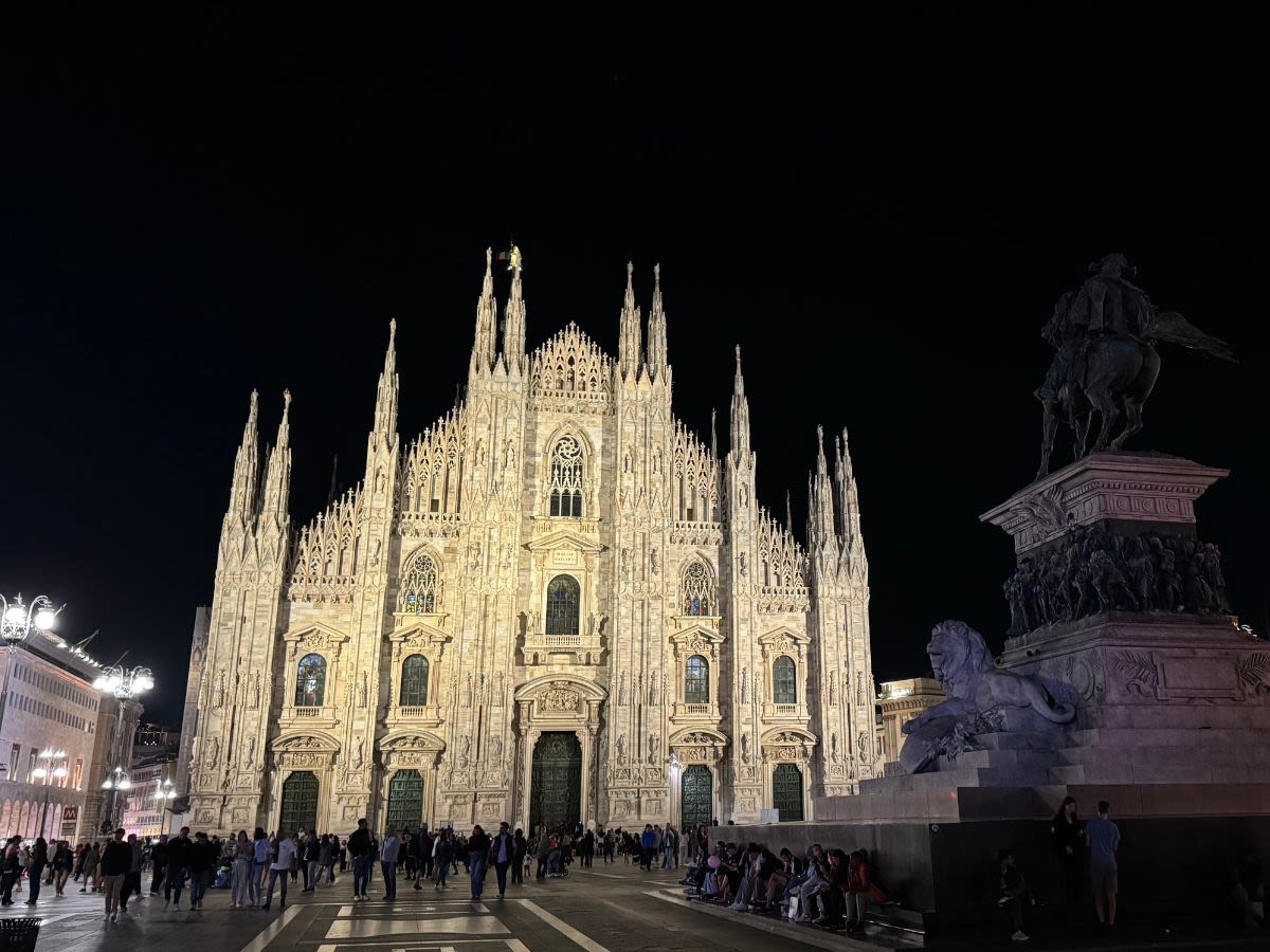 Katedra Duomo di Milano w piątkowy wieczór. Oświetlona fasada budynku kontrastuje z czarnym niebiem. Przed katedrą na placu przechadzają się turyści.