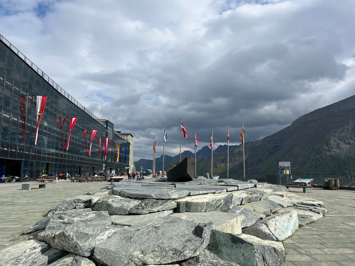 Plac przed parkingiem przy lodowcu Pasterze na trasie Grossglockner Hochalpenstrasse. Na placu znajdują się kamienne głazy oraz drewniana łódź. Turyści chodzą po placu. 