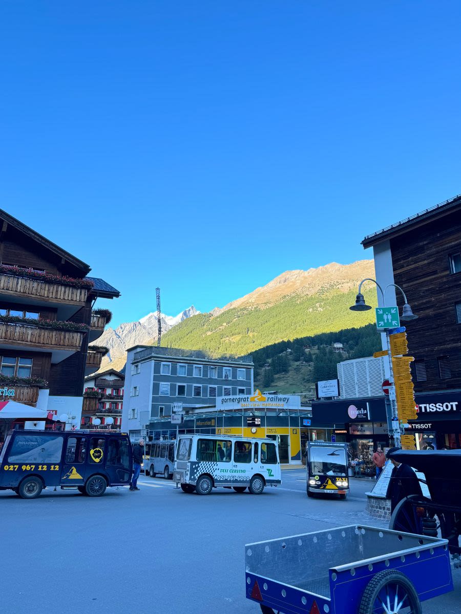 Zermatt, górskie miasteczko, do którego przyjeżdżają osoby, które chcą zobaczyć Matterhorn. Widać drewniane budynki i kilka pojazdów elektrycznych, które wożą turystów.