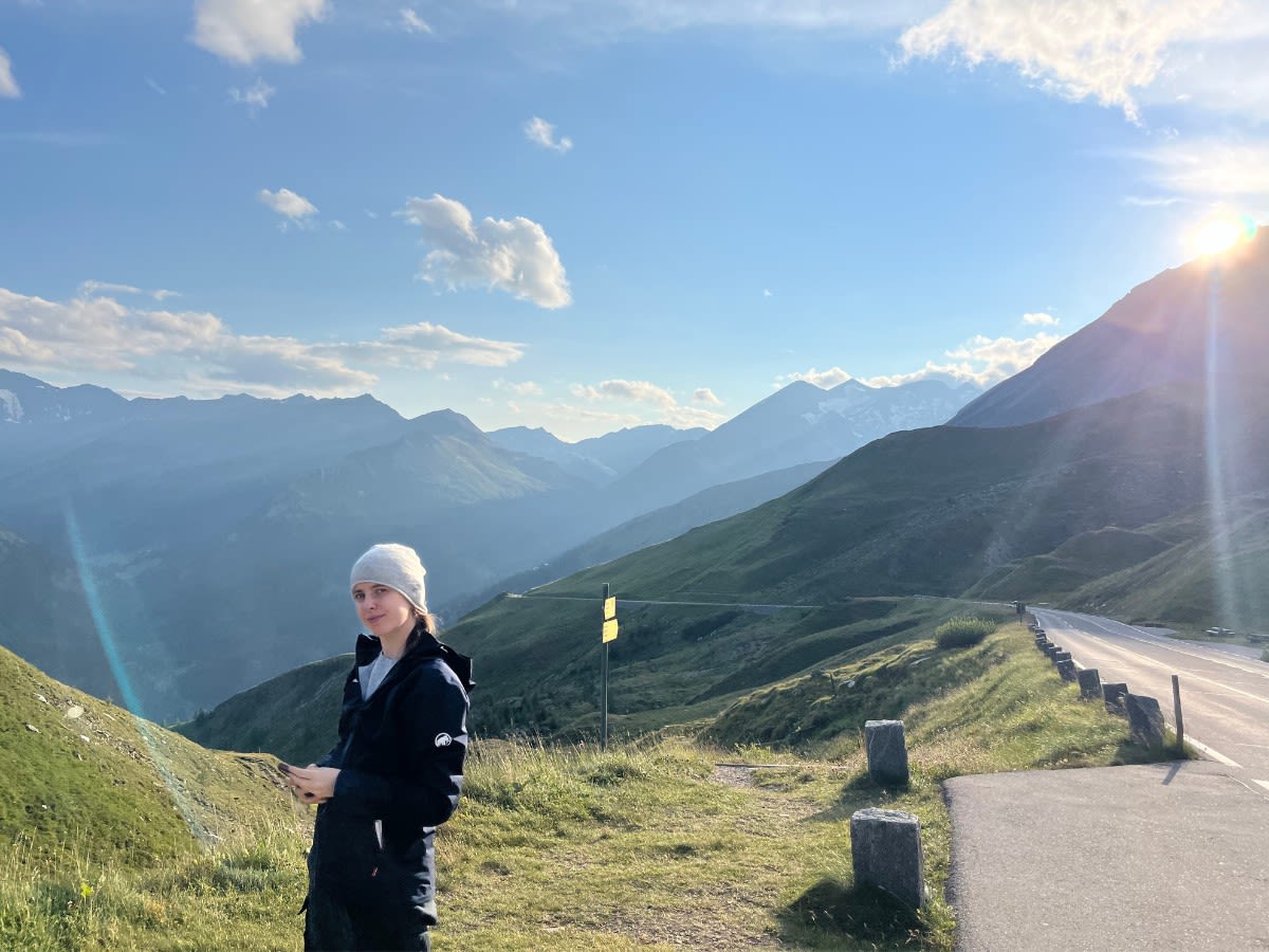 Autorka bloga Słowa Płyną stoi przy alpejskiej drodze Grossglockner Hochalpenstrasse. W tle panorama gór i słońce, które zachodzi za górami. 