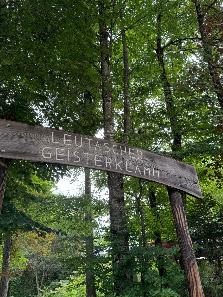 Na drewnianej tablicy widnieje napis Leutascher Geisterklamm. Jest do wejście do wąwozu Leutascher Geisterklamm w Austrii. Wzdłuż ścieżki rosną wysokie iglaste drzewa.