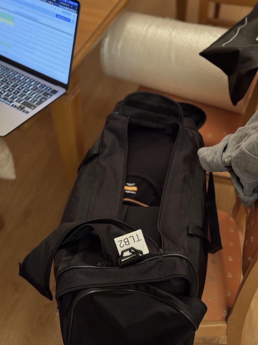 Czarna torba jest otwarta a w niej ubrania. Do torby przyczepiona jest karteczka z legendą. Torba leży na krześle. Obok na stole leży laptop.