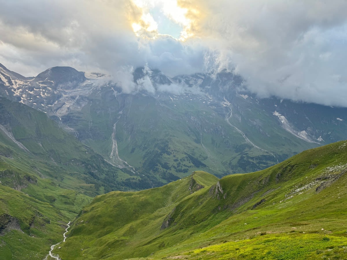 Widoki z alpejskiej drogi Grossglockner Hochalpenstrasse w lecie. Niższe partie gór pokryte są trawą o intensywnym kolorze. Wyższe śniegiem lub są nagie. W dole płynie rzeka.