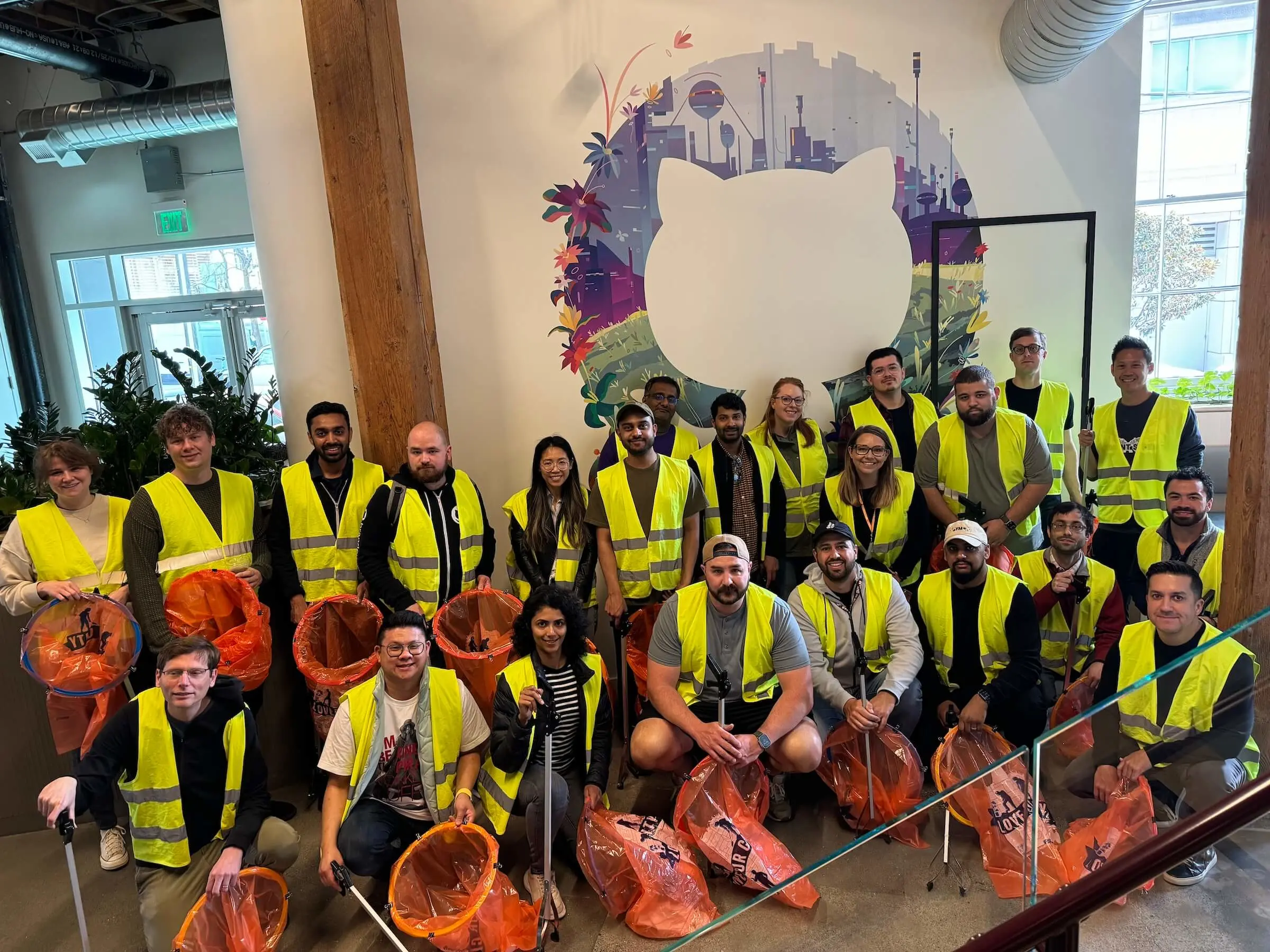 A large group of people wearing yellow safety vests is gathered indoors in front of a wall featuring the GitHub logo. They are holding orange trash bags and litter pickers, suggesting they are participating in a cleanup activity. 