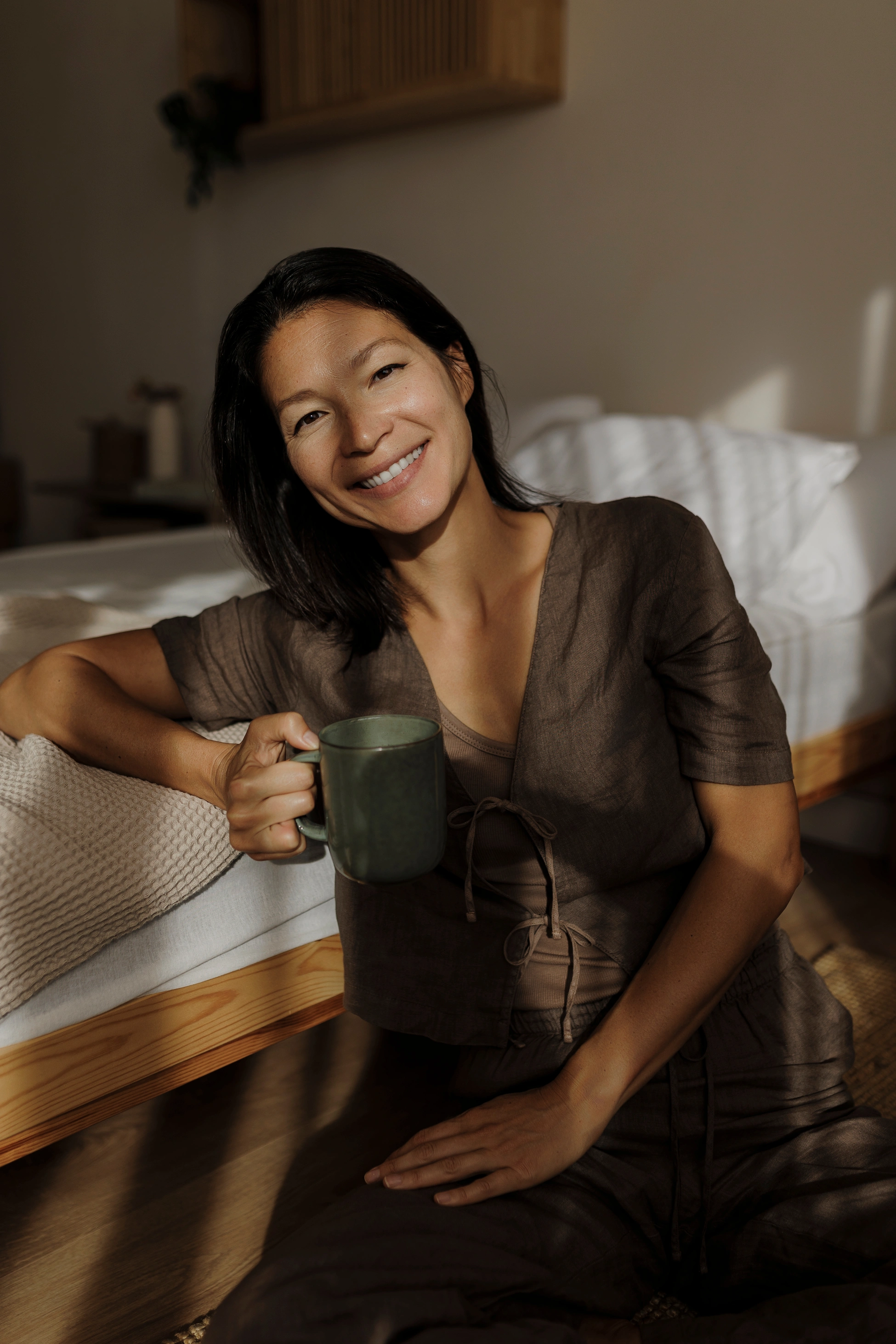 Eine Frau im Pyjama sitzt lächelnd auf dem Boden ihres Schlafzimmers und hält eine Tasse Kaffee in der Hand.