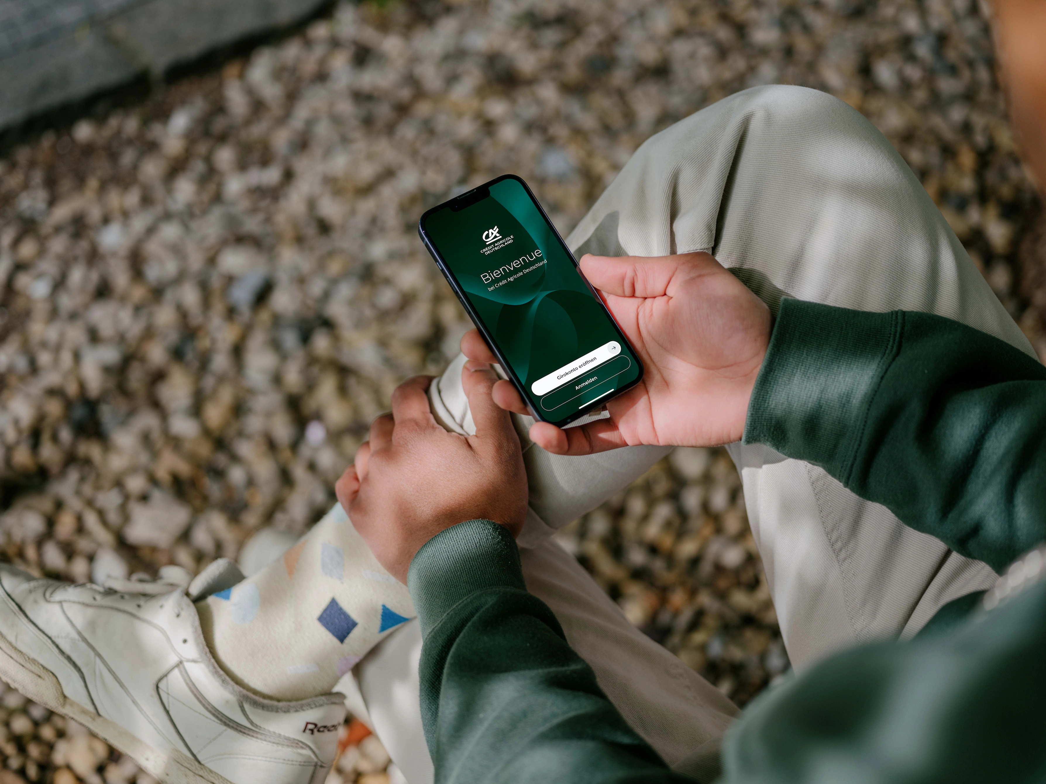 Eine Person in grünem Pullover und heller Hose sitzt im Freien auf Kies und hält ein Smartphone in der Hand. Auf dem Bildschirm ist der Startbildschirm der Banking-App zu sehen, mit der Begrüßung „Bienvenue bei Crédit Agricole Deutschland“ und den Schaltflächen „Girokonto eröffnen“ und „Anmelden“.