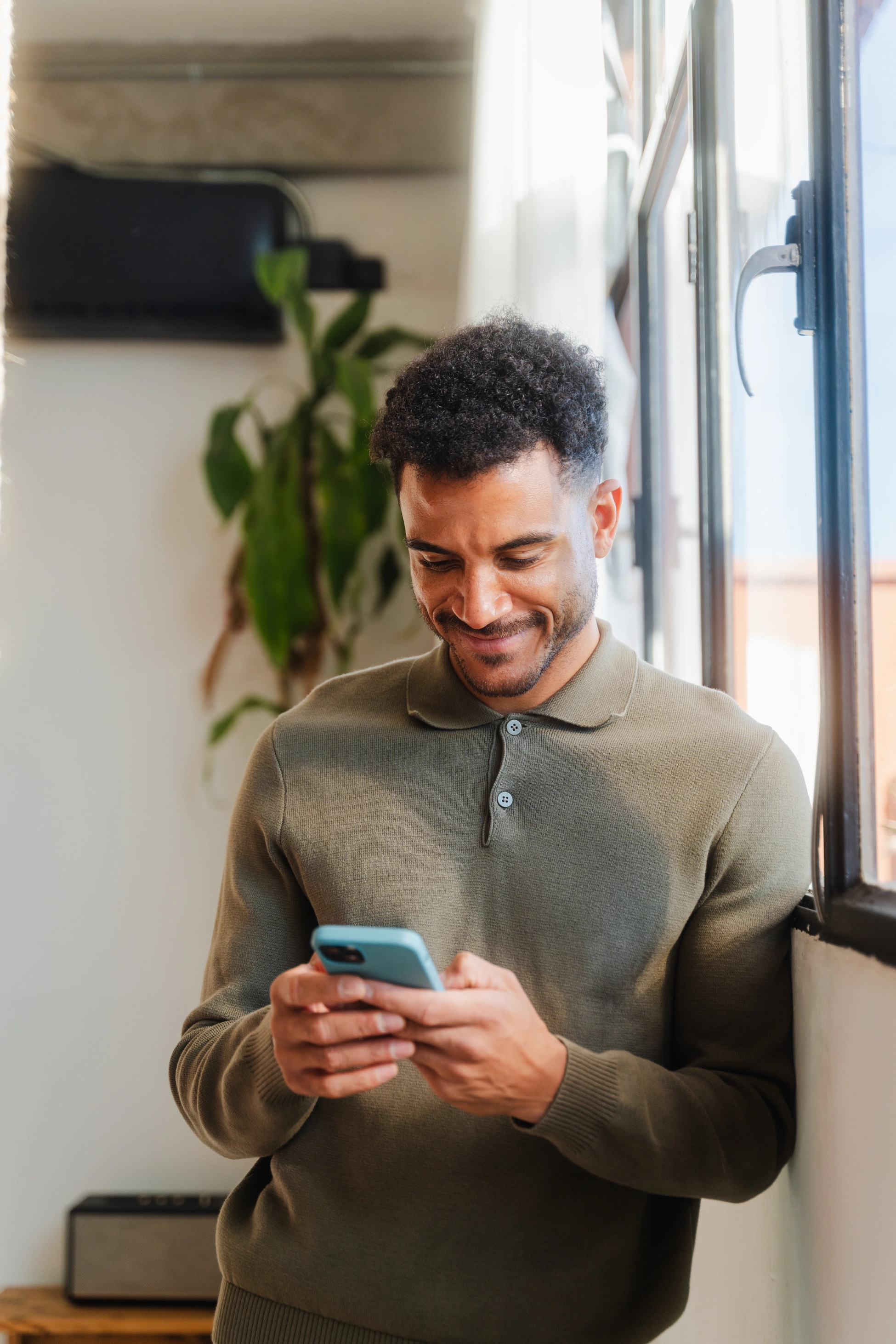 Porträt eines jungen Mannes in Smart-Casual-Kleidung, der im Büro am Fenster lehnt und Nachrichten auf seinem Smartphone schreibt