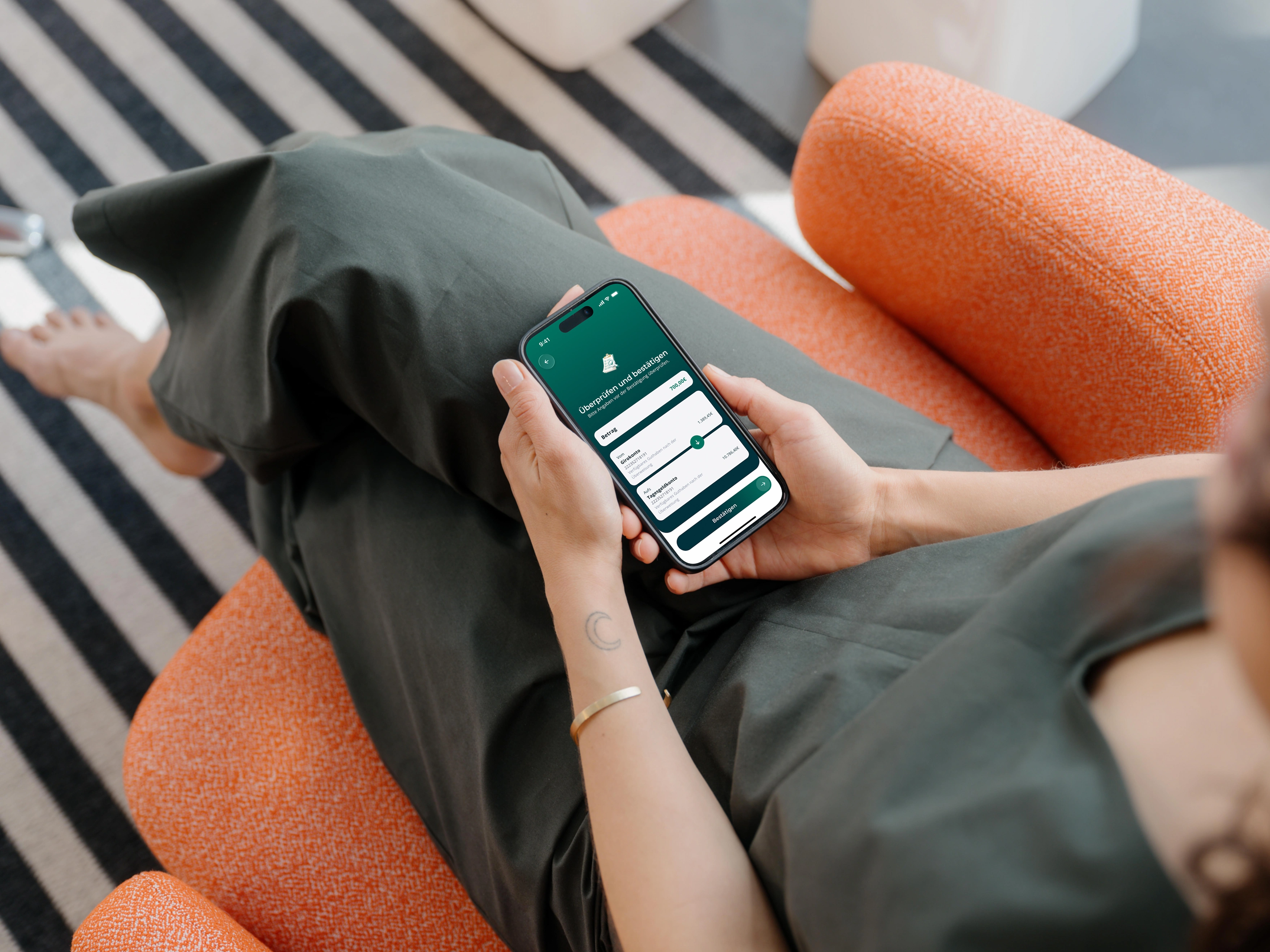 Eine Frau in einem dunkelgrünen Kleid sitzt mit übergeschlagenen Beinen in einem orangefarbenen Sessel und hält ein Smartphone in beiden Händen. In der Banking-App prüft sie eine Umbuchung von 700 € vom Girokonto auf das Tagesgeldkonto und bestätigt sie. Am Boden liegt ein schwarz-weiß gestreifter Teppich.