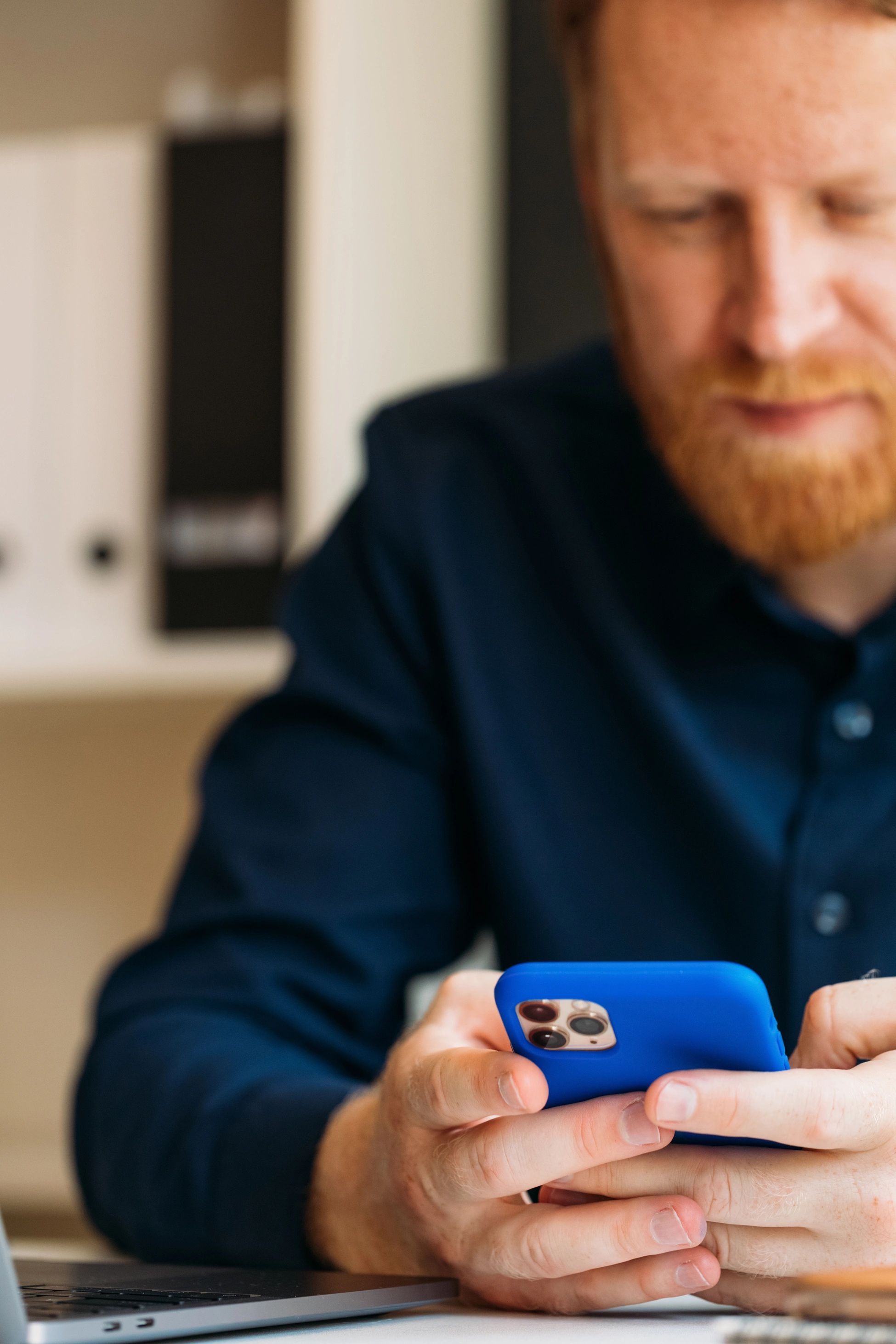Rothaariger Mann mit Bart in einem dunkelblauen Hemd sitzt im Büro am Schreibtisch und nutzt sein Smartphone.