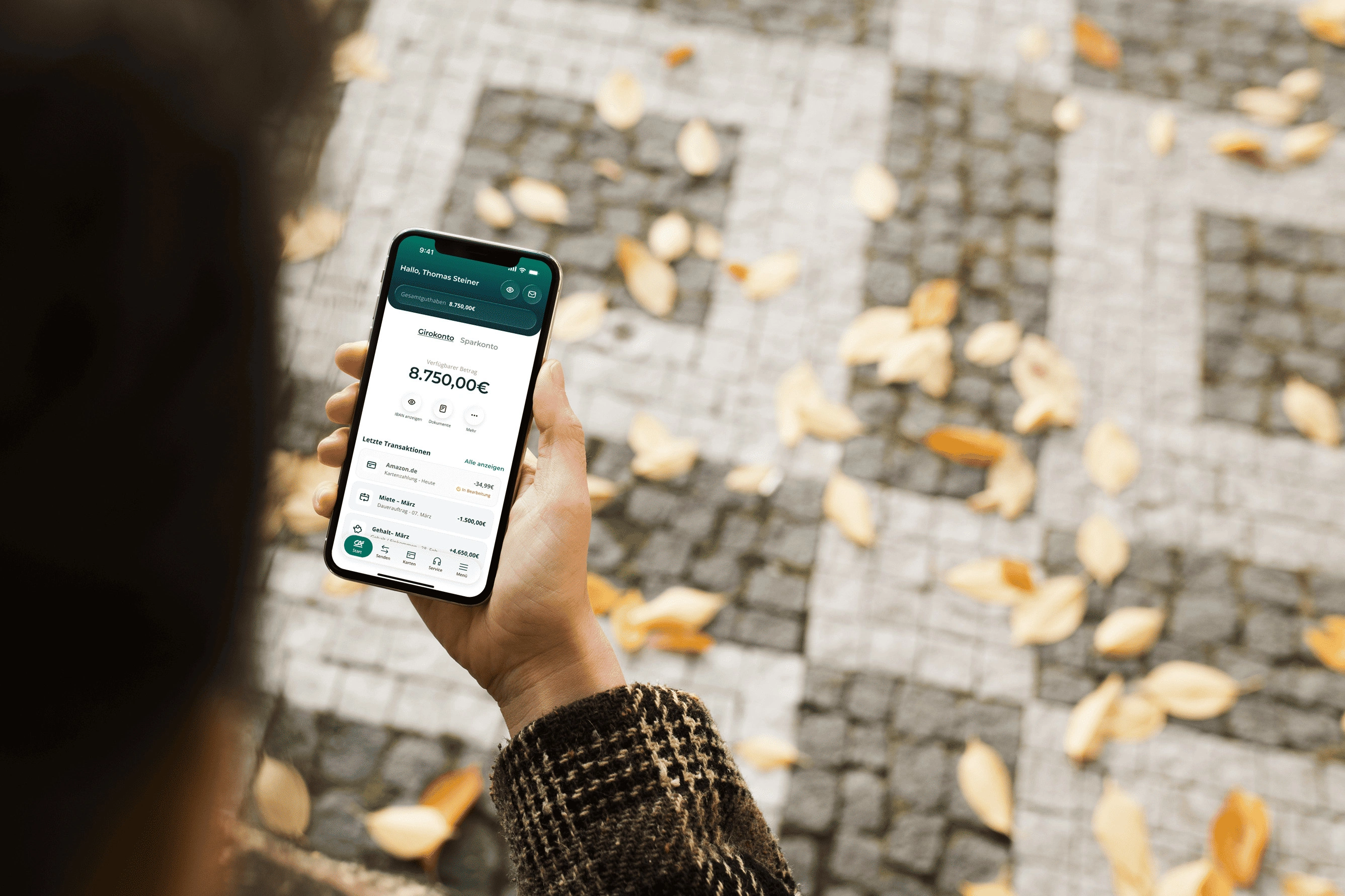 Eine Person in einer gemusterten Strickjacke hält ein Smartphone mit geöffneter Banking-App in der Hand. Auf dem Bildschirm sind der Kontostand und die letzten Transaktionen des Girokontos zu sehen. Im Hintergrund liegt Herbstlaub auf grau-weissem Kopfsteinpflaster.