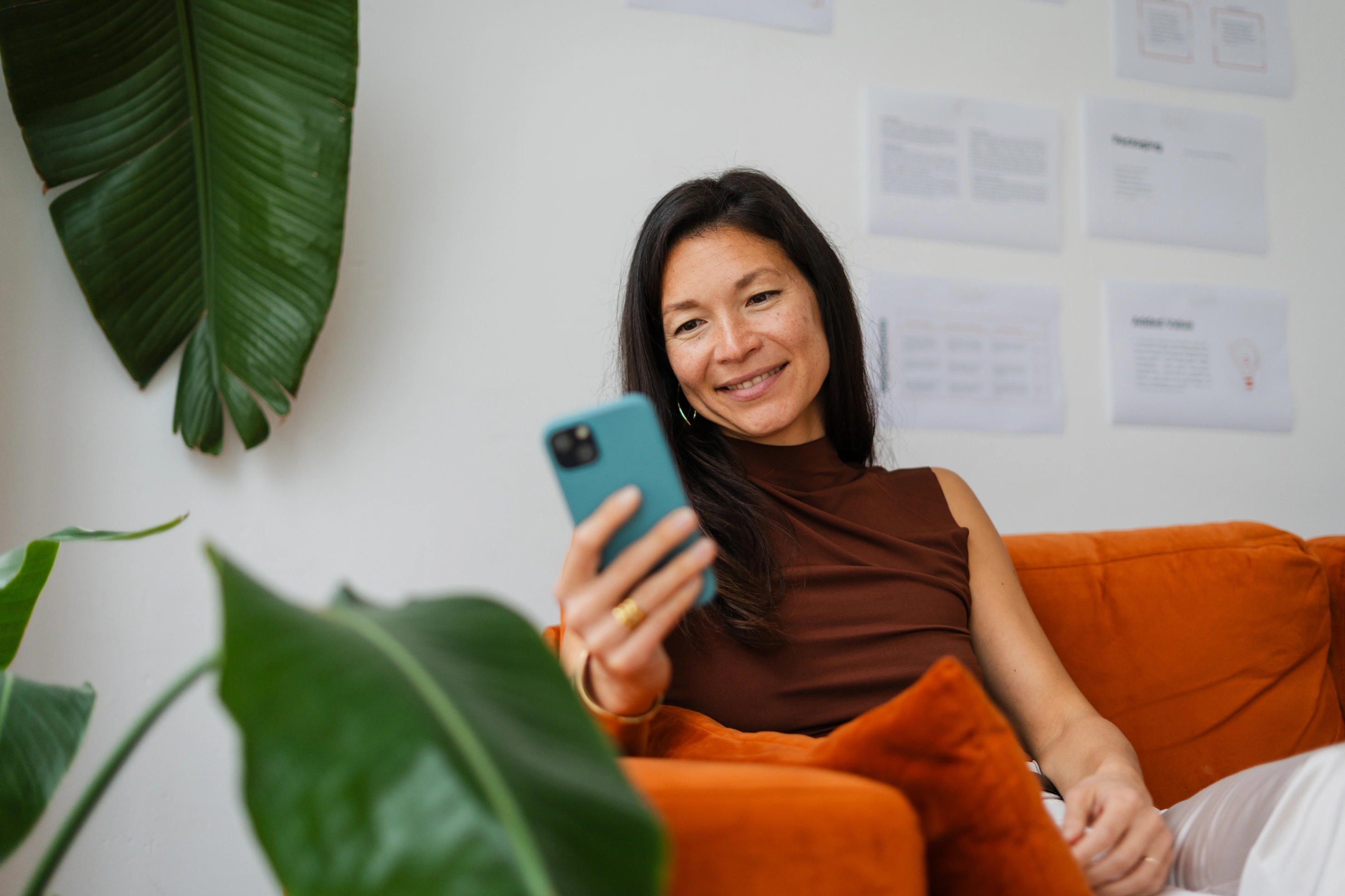 Eine Geschäftsfrau sitzt in der Lounge-Ecke des Büros auf einem leuchtend orangefarbenen Sofa und lächelt, während sie ihr Smartphone in der Hand hält.
