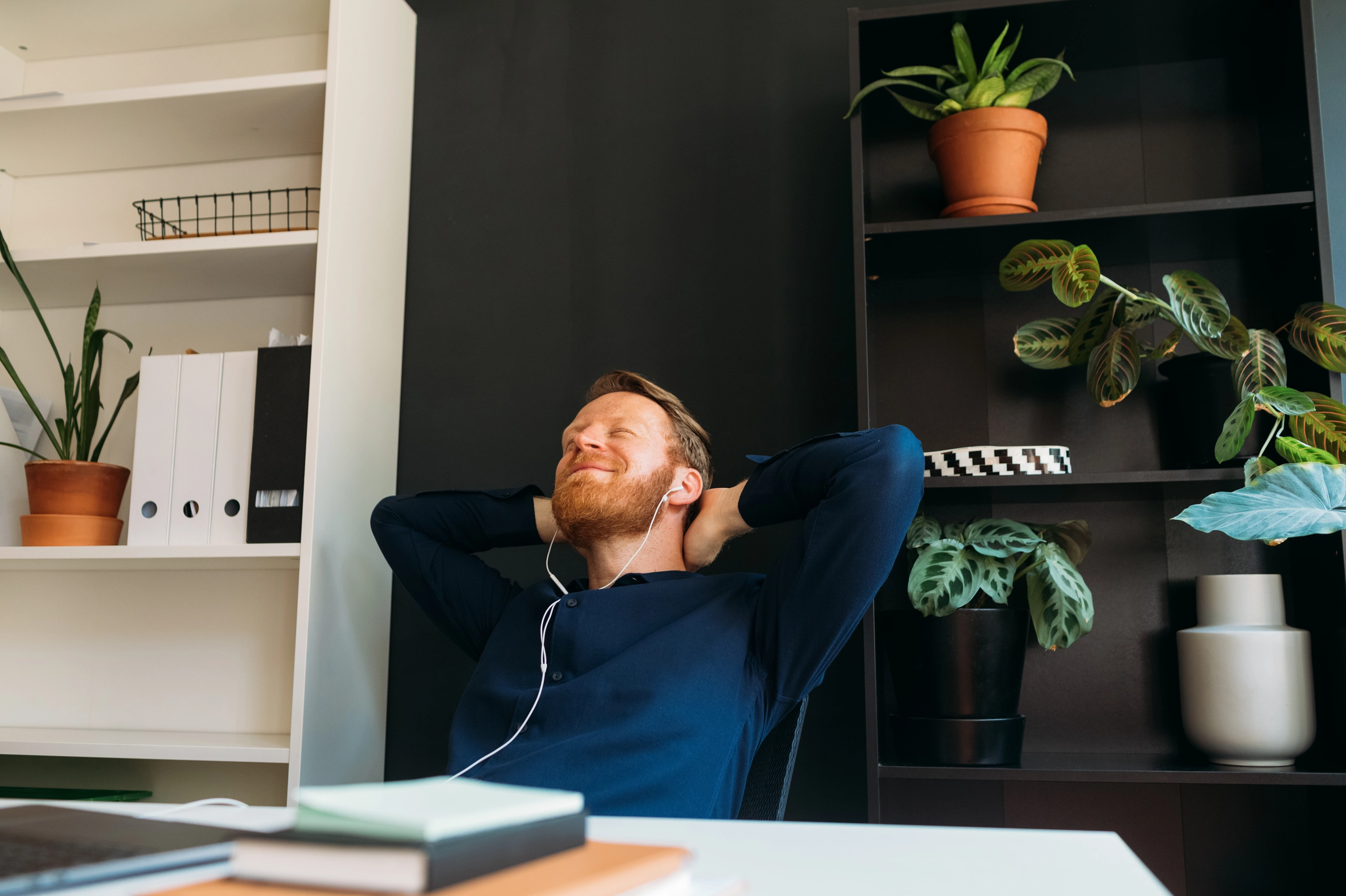 Porträt eines rothaarigen Mannes mit In-Ear-Kopfhörern, der mit geschlossenen Augen im Büro sitzt, entspannt und Musik hört.