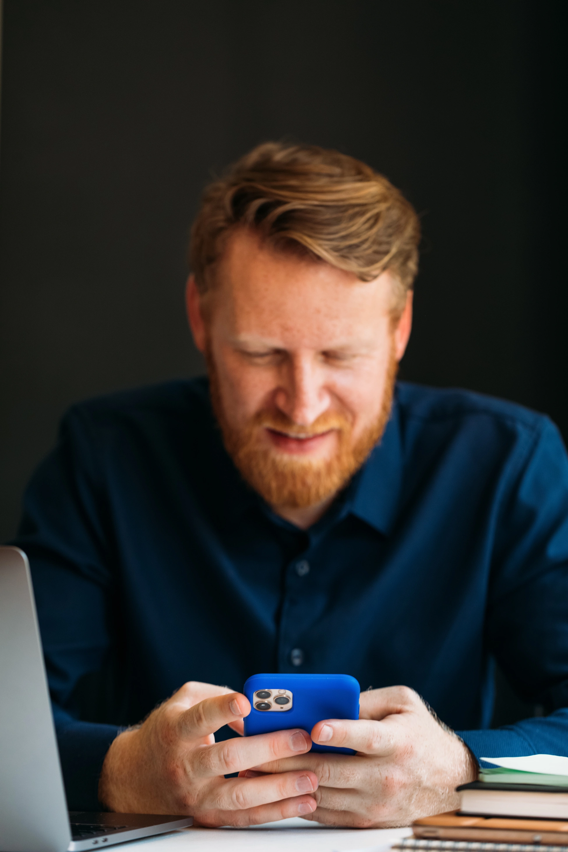 Ein Mann mit Bart und rotem Haar trägt ein dunkelblaues Hemd. Er sitzt an seinem Schreibtisch und ist mit seinem Smartphone beschäftigt.