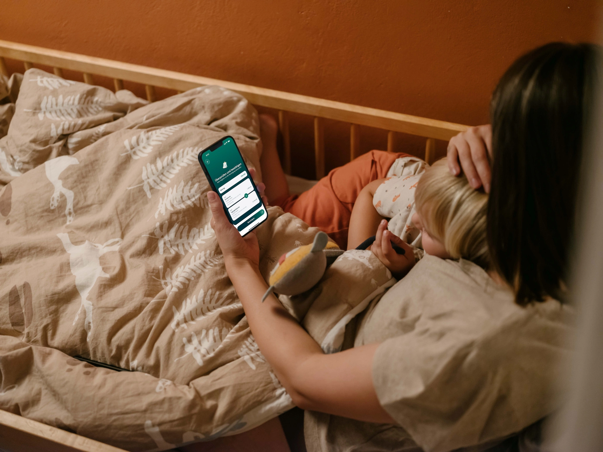 Eine Frau liegt mit einem kleinen Kind im Kinderbett und streichelt ihm über den Kopf. In der anderen Hand hält sie ein Smartphone mit geöffneter Banking-App. Auf dem Bildschirm prüft sie eine Umbuchung vom Girokonto auf das Tagesgeldkonto und bestätigt sie. Im Hintergrund ist eine warme, orangefarbene Wand zu sehen.